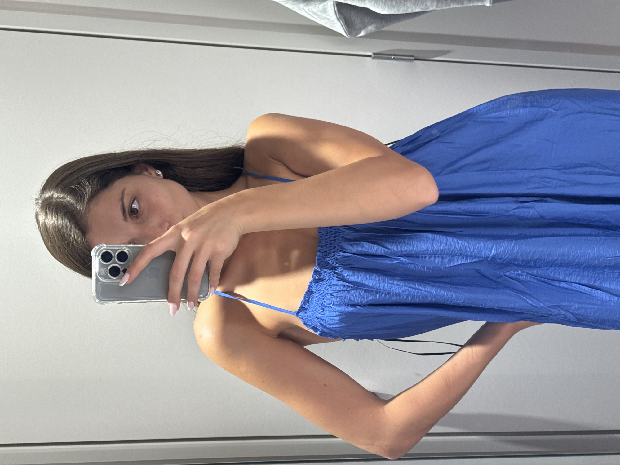 In a fitting room, a young woman with long brown hair takes a selfie in a mirror. She is wearing a bright blue spaghetti strap dress with a gathered bodice. Her left hand is on her hip, while her right hand holds a silver iPhone with a clear case, obscuring part of her face. Her nails are painted a pale pink. The background is a plain, light grey wall, characteristic of a retail fitting room. There are vertical lines suggesting doors or panels, and to the right, a grey hooded sweatshirt is visible, hanging on a hook. The lighting is bright and even, casting soft shadows. The location is described as a fitting room in Burgas, Bulgaria.