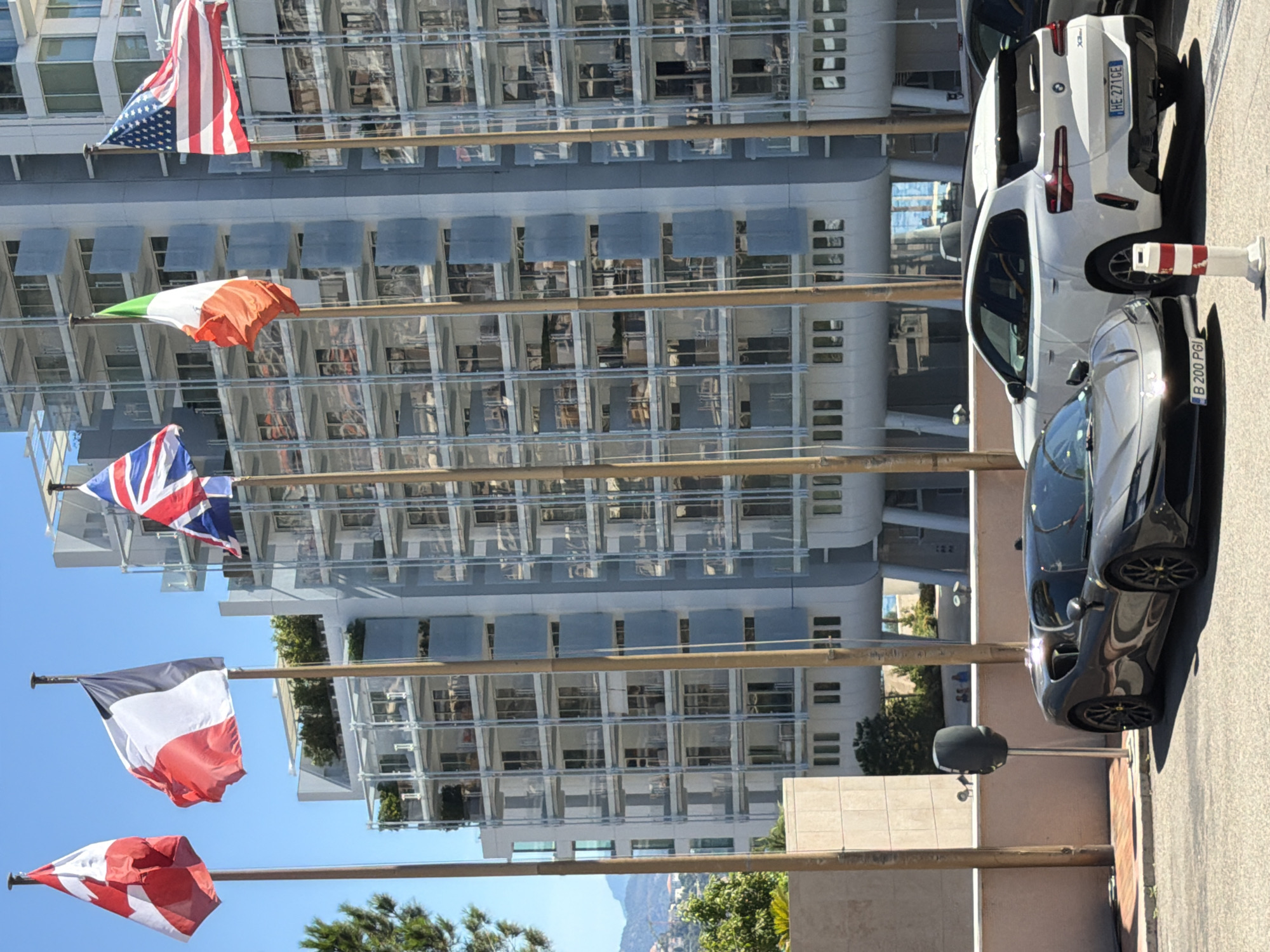 The image depicts a street scene in Monaco, with a modern building as the backdrop. In the foreground, a dark gray sports car is parked on the right side of the frame, facing left. To its right, a white SUV is also parked. Several flags are displayed on poles in front of the building: the Canadian flag on the far left, followed by the French flag, the Union Jack, the Italian flag, and the American flag on the far right. The building itself is tall and modern, with multiple floors featuring balconies and glass facades. The sky is clear and blue, suggesting a sunny day.