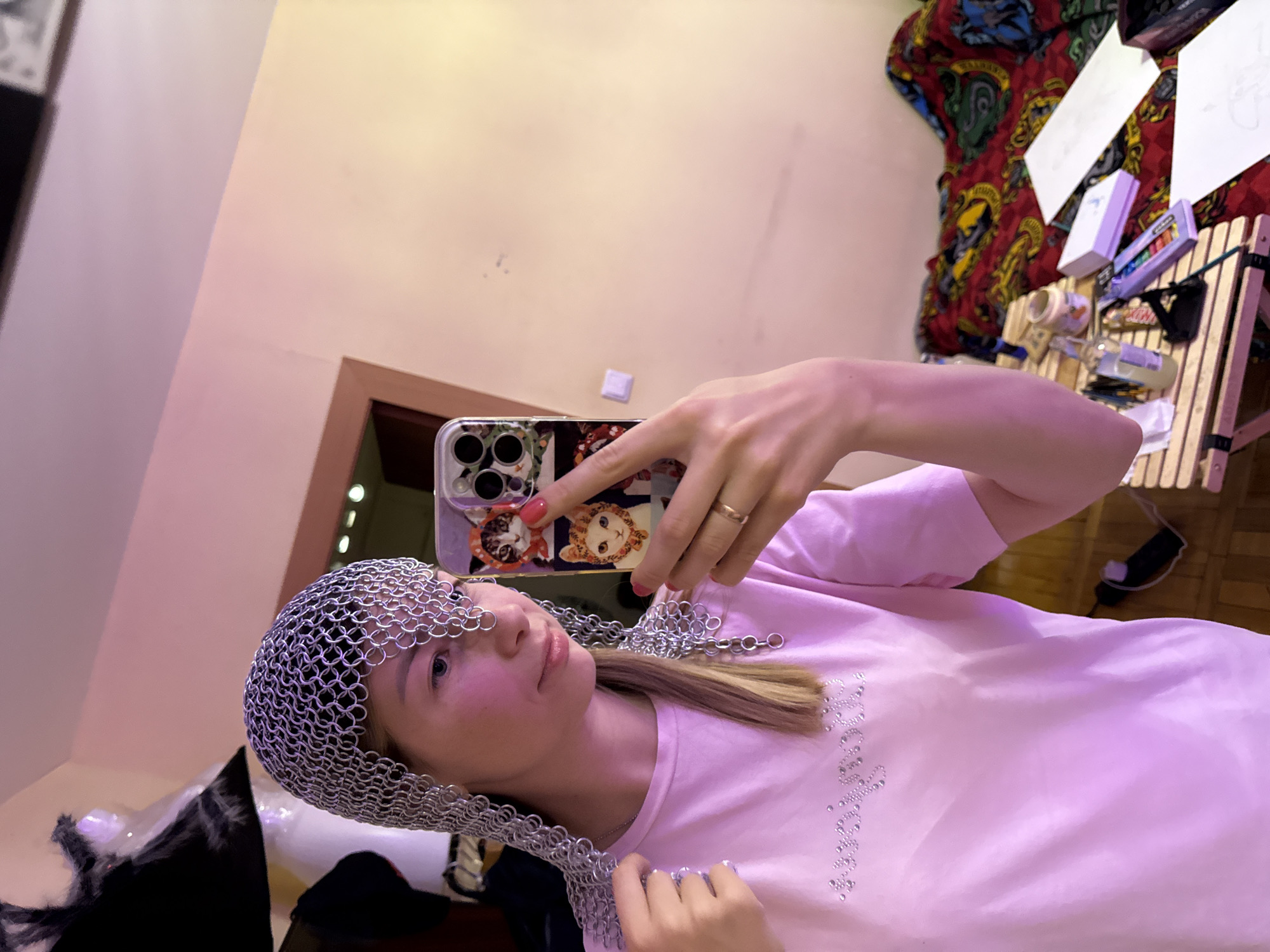 A young female, with light skin and light brown hair partially visible, is depicted indoors in what appears to be a room in Moscow, Russia. She is taking a mirror selfie, holding a silver-colored smartphone with a multi-lens camera system in her left hand. The phone is encased in a purple-toned cover featuring cartoon images of cats wearing headwear. A gold-colored ring is present on the ring finger of her left hand, and her fingernails are painted pink.
She is wearing a short-sleeved, light pink t-shirt with a sparkling rhinestone embellishment across the chest that spells out a word or phrase, partially visible. She is also wearing a piece of chainmail on her head, which extends to partially cover her forehead, right temple, and drapes over her shoulders. Her right hand is lightly touching the chainmail on her right shoulder. Her expression is a slight, closed-mouth smile.
The background wall is a light, warm tone, possibly beige or pale yellow, with a prominent pink-purple hue cast upon the upper wall and ceiling, indicating artificial colored lighting. To the left, a dark door frame and some dark fabric on a hanger or stand are visible. To the right, a bed is present, covered with a patterned bedspread in red, yellow, blue, and green hues, featuring an intricate ornamental design. A small, slatted wooden table is positioned in front of the bed, holding various items: a clear glass bottle with light liquid, a small light-colored cylindrical object, a purple rectangular box, a small dark object, several sheets of white paper with visible pencil sketches, and a dark rectangular box. A black cable is visible near the table and bed.