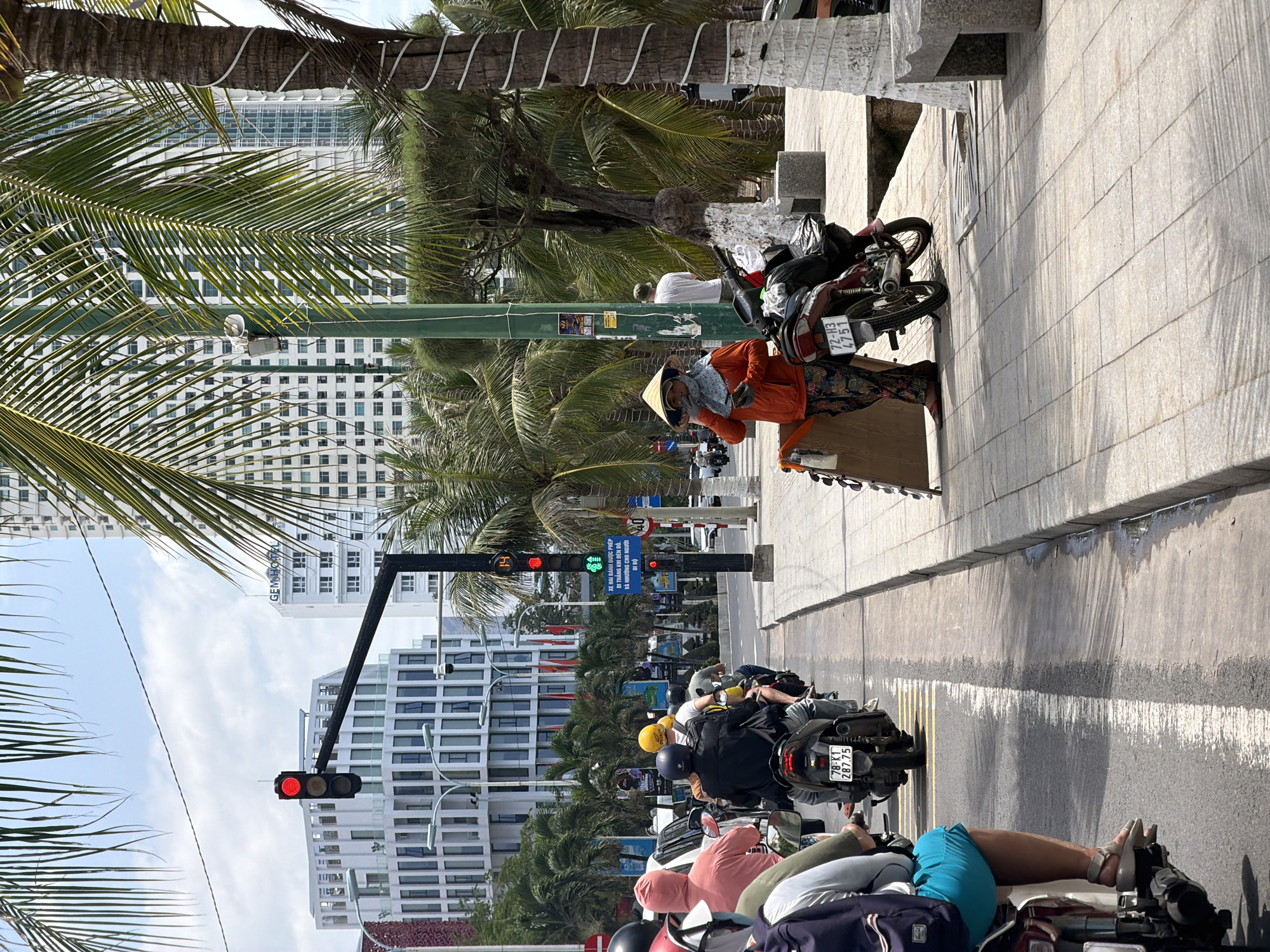 This media file presents a bustling daytime street scene in Nha Trang, Vietnam, characterized by urban activity under a bright, partly cloudy sky. The image captures a street lined with modern, multi-story buildings and numerous palm trees, giving it a tropical city atmosphere.
On the right side, a paved sidewalk features a person wearing an orange long-sleeved shirt and a traditional Vietnamese conical hat (nón lá), standing next to a parked moped with the license plate "72-H3 4751". This individual appears to be a vendor or someone taking a break. Further back on the sidewalk, another person in a white t-shirt is partially visible.
The street is busy with numerous mopeds and scooters, forming a queue at a red traffic light in the foreground. Riders, most wearing helmets, patiently await the signal change. One moped in the middle ground displays the license plate "78-K1 287.75". A digital countdown timer on the traffic light shows "39", indicating the seconds remaining before the light turns green. Further down the street, another traffic light appears to be green.
Notable textual details include "GEM HOT" on a large building in the background, likely part of a hotel name. A prominent sign on a green pole carries Vietnamese text: "XE HAI BÁNH ĐƯỢC PHÉP ĐI THẲNG KHI ĐÈN ĐỎ. VÀ NHƯỜNG CHO NGƯỜI ĐI BỘ," which translates to "TWO-WHEELED VEHICLES ARE ALLOWED TO GO STRAIGHT WHEN THE RED LIGHT IS ON. AND YIELD TO PEDESTRIANS." A speed limit sign indicating "40" is also visible. The scene offers a glimpse into the daily life and infrastructure of a Vietnamese city.