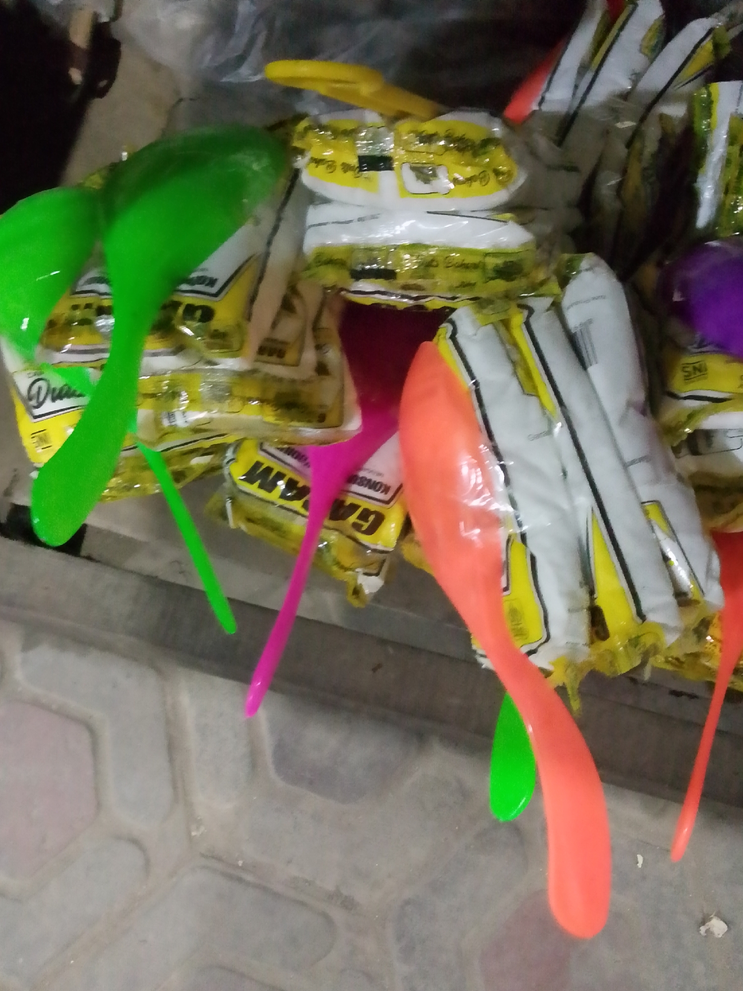 The image shows a close-up, overhead view of colorful plastic ladles (sendok sayur) arranged on a paved surface, likely in Bulurejo, Indonesia. Several green, pink, and orange ladles are visible, some partially obscured by packaging. The packaging appears to be for consumer goods, with yellow and white labels featuring text in Indonesian. The background is dark and out of focus, suggesting an indoor setting like a shop or market stall. The paved surface is a light gray color with a patterned texture, resembling interlocking pavers. There are no people visible in the image, and the focus is entirely on the merchandise. The lighting is dim, with some reflections visible on the plastic surfaces.