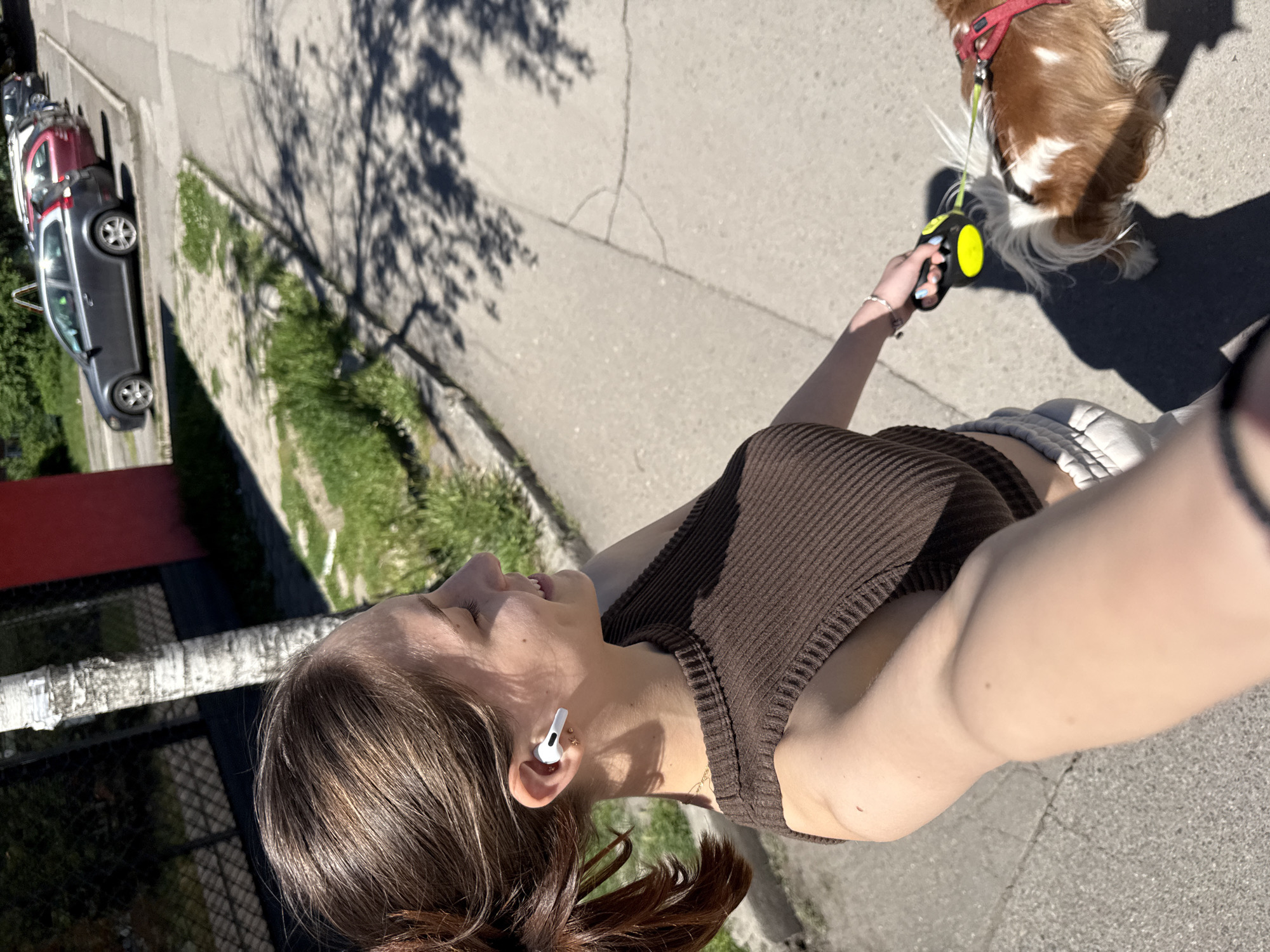 A young woman is taking a selfie while walking a brown and white dog on a leash. The woman is wearing a brown tank top and white earbuds. In the background, there are parked cars, greenery, and the shadow of a tree. The location is likely a residential street with sidewalks and trees, potentially in Sofia, Bulgaria.