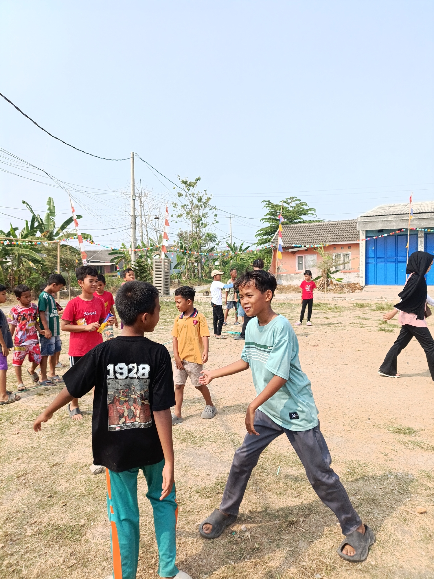 A group of children are gathered outdoors in what appears to be a village or rural area in Cariu, Indonesia. The ground is a mixture of dry grass and dirt. In the background, there are houses with blue doors, and trees with banana leaves. Several flags, likely red and white, are visible, indicating a possible celebration or event. The children are engaged in various activities, with some appearing to be playing or interacting with each other. One boy in the foreground is wearing a black t-shirt with the number "1928" printed on the back and teal pants with orange stripes. Another boy is wearing a light blue t-shirt and grey pants. A girl in a black hijab and pink shirt is walking in the background. The sky is clear and blue.