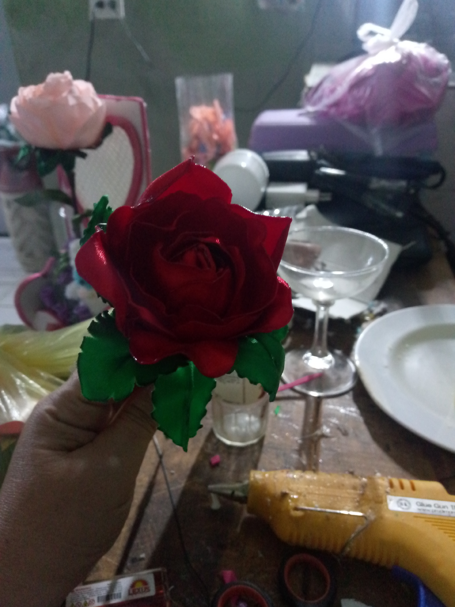 A close-up shot depicts a person holding a red rose with green leaves. The background is cluttered with various objects including a champagne glass, a white plate, a glue gun, spools of tape, and other crafting materials. A pink rose can also be seen in the background, along with a heart-shaped mirror frame. The setting appears to be a crafting area. The location is stated as Juan Griego, Venezuela.
