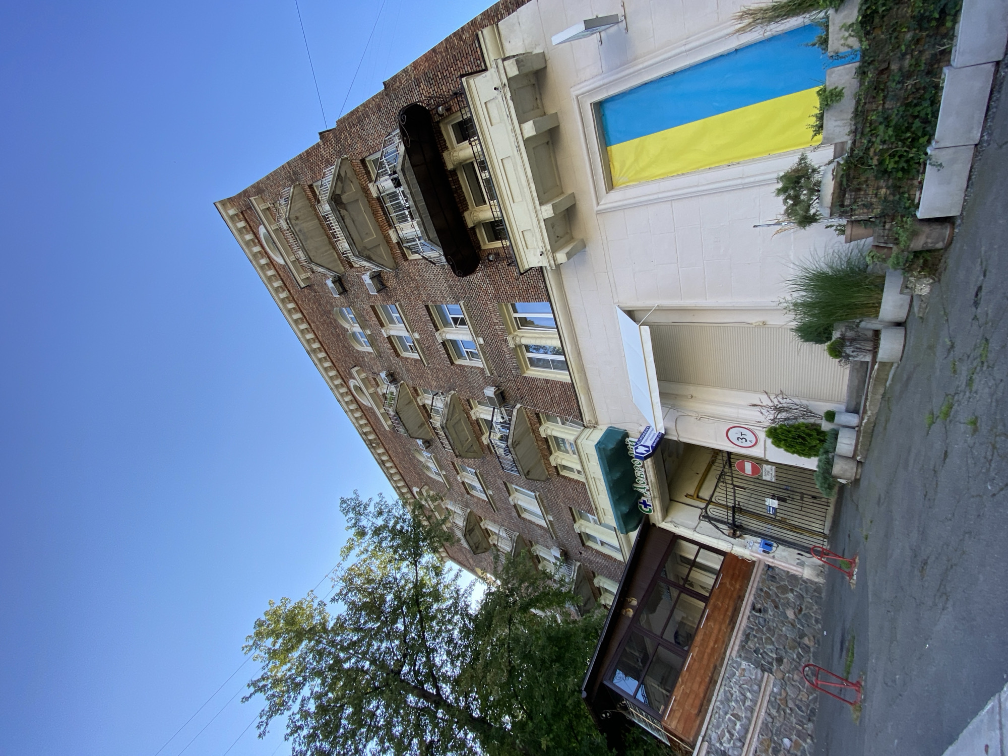 A low-angle, wide shot captures the facade of a brick apartment building under a clear blue sky. The building features numerous balconies with white railings and some have black awnings or decorative elements. Green trees frame the left side of the building, with some branches extending over the structure. On the lower part of the building, a large Ukrainian flag is displayed vertically in a window or alcove, with its blue and yellow colors prominently visible. Several potted plants and bushes are arranged in front of the building, some on a stone retaining wall and others in concrete planters. A closed gate with signage and a red circle with a "3t" inside is visible in the middle of the frame, suggesting a restricted area or entrance. A small wooden structure with large windows is attached to the left side of the building, appearing to be a cafe or extension. Red bollards are present on the asphalt ground in the foreground. The scene depicts an urban setting with residential architecture and patriotic symbolism.