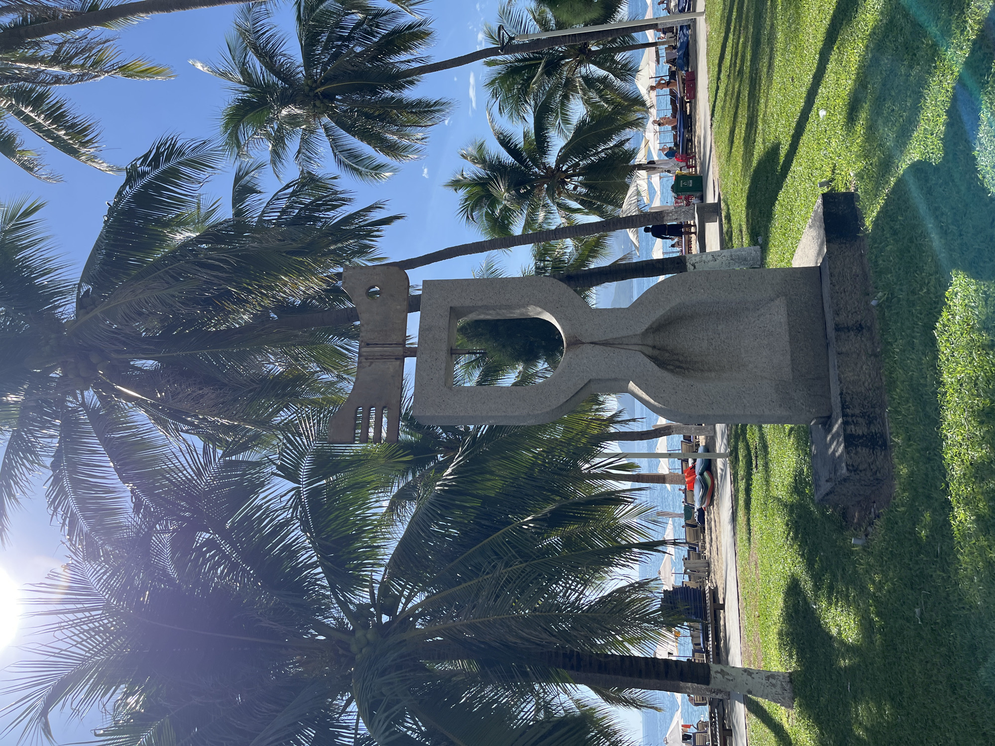 This image depicts a sculpture made of granite, located in a grassy area by the beach in Nha Trang, Vietnam. The sculpture is abstract, with a large, curved base and an upper section resembling a stylized key or symbol. It is surrounded by lush green grass, with palm trees casting shadows across the lawn.
In the background, the turquoise waters of the sea stretch towards the horizon under a clear blue sky. A beach lined with lounge chairs and white umbrellas is visible, with several people scattered along the shoreline. Some individuals are seen walking or relaxing on the beach, while others are partially obscured by the palm trees. The overall scene evokes a tranquil and sunny atmosphere, characteristic of a tropical coastal city. The presence of the sculpture suggests it is a public art installation, likely a landmark or a decorative element in a park or promenade area.
