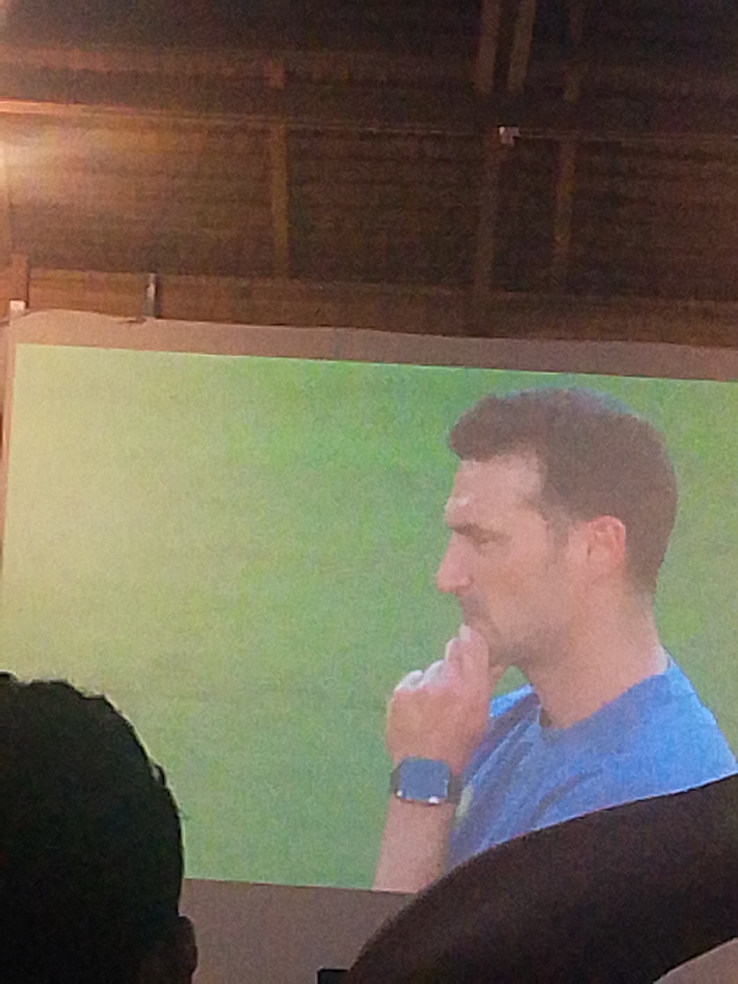The image shows a man in profile, wearing a blue shirt and a smartwatch on his left wrist. He has his hand on his chin, appearing to be in thought. The background is a blurry green, possibly a field or a screen displaying an image. In the foreground, a dark, blurry shape, likely the back of someone's head, partially obstructs the view. The overall lighting is dim and appears to be artificial. The image is grainy and low resolution, suggesting it was taken in low light conditions or from a distance. There is no visible indication of the specific location within Meunasahmee, Indonesia.