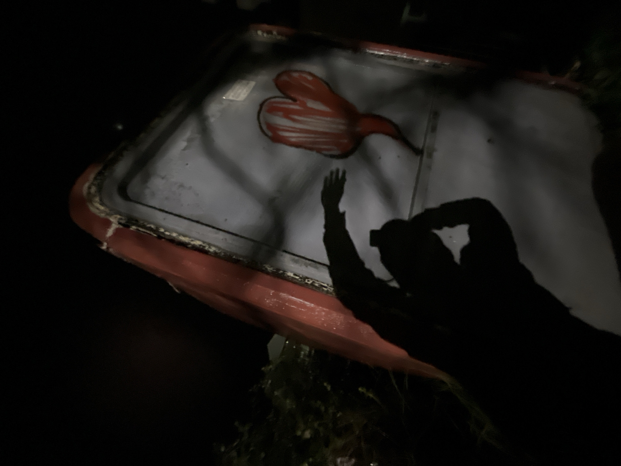 The scene is set outdoors at night in Budva, Montenegro. A large, overturned, red-orange fiberglass object, possibly a boat hull or a substantial container, occupies the central and right portions of the frame. Its exposed greyish interior or underside serves as a canvas for graffiti, which depicts a red, somewhat irregular heart-like or mitten shape with vertical white stripes and a dark outline. A prominent, dark human shadow is cast across the lower-left and central grey surface, showing an individual holding a rectangular device, consistent with a smartphone, with both hands, in the act of photographing the graffiti. Dark vegetation is visible to the left and beneath the overturned object, and a faintly illuminated building structure can be discerned in the distant background.