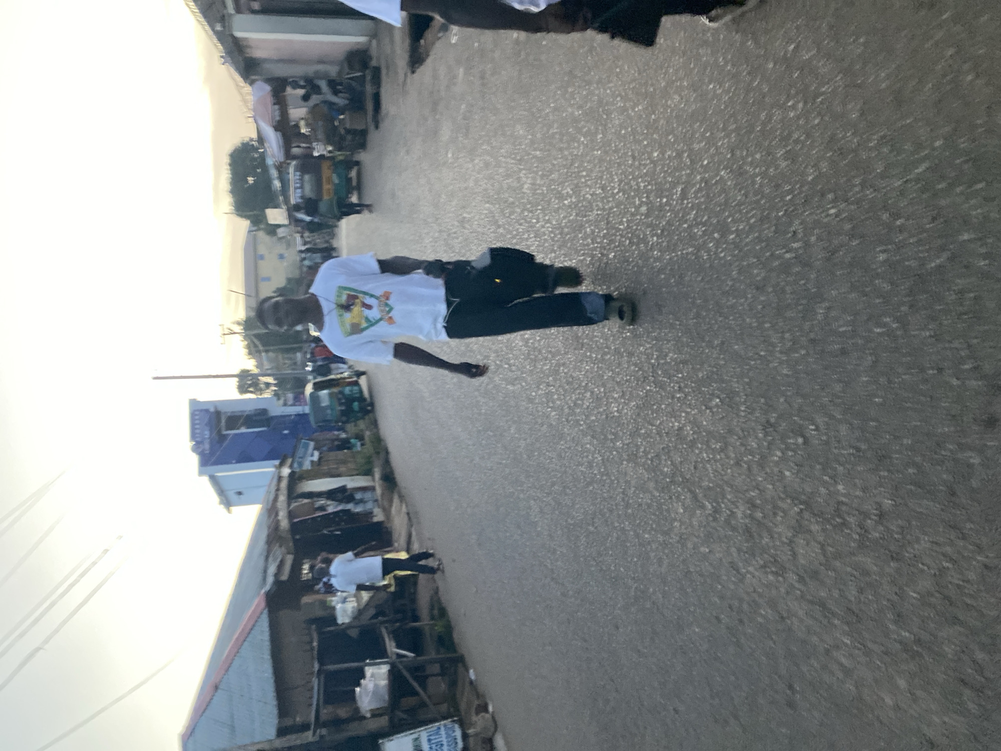 A man in a white t-shirt with a graphic design and dark pants walks down a paved street in Jos, Nigeria. He carries a dark bag in his right hand. The street is lined with buildings, some appearing to be shops, with corrugated metal roofs. A green auto-rickshaw is parked on the right side of the street. In the background, several other people are visible, and the sky is bright with what appears to be a sunrise or sunset. The overall scene depicts a typical urban street environment in Jos.
