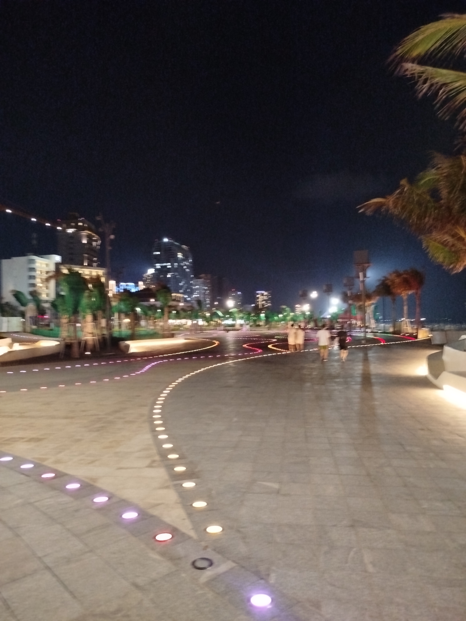 At night, the image depicts a wide, paved walkway, likely a promenade or public space in the city of Ho Chi Minh City, Vietnam. The walkway curves gently and is lined with inset lights that emit colorful glows, including purple, pink, and yellow hues. The city skyline, featuring multiple illuminated buildings, is visible in the background. There are a few people walking along the walkway. To the right of the image, parts of palm trees are visible. The lighting is a mix of ambient city light and the artificial lights from the walkway.