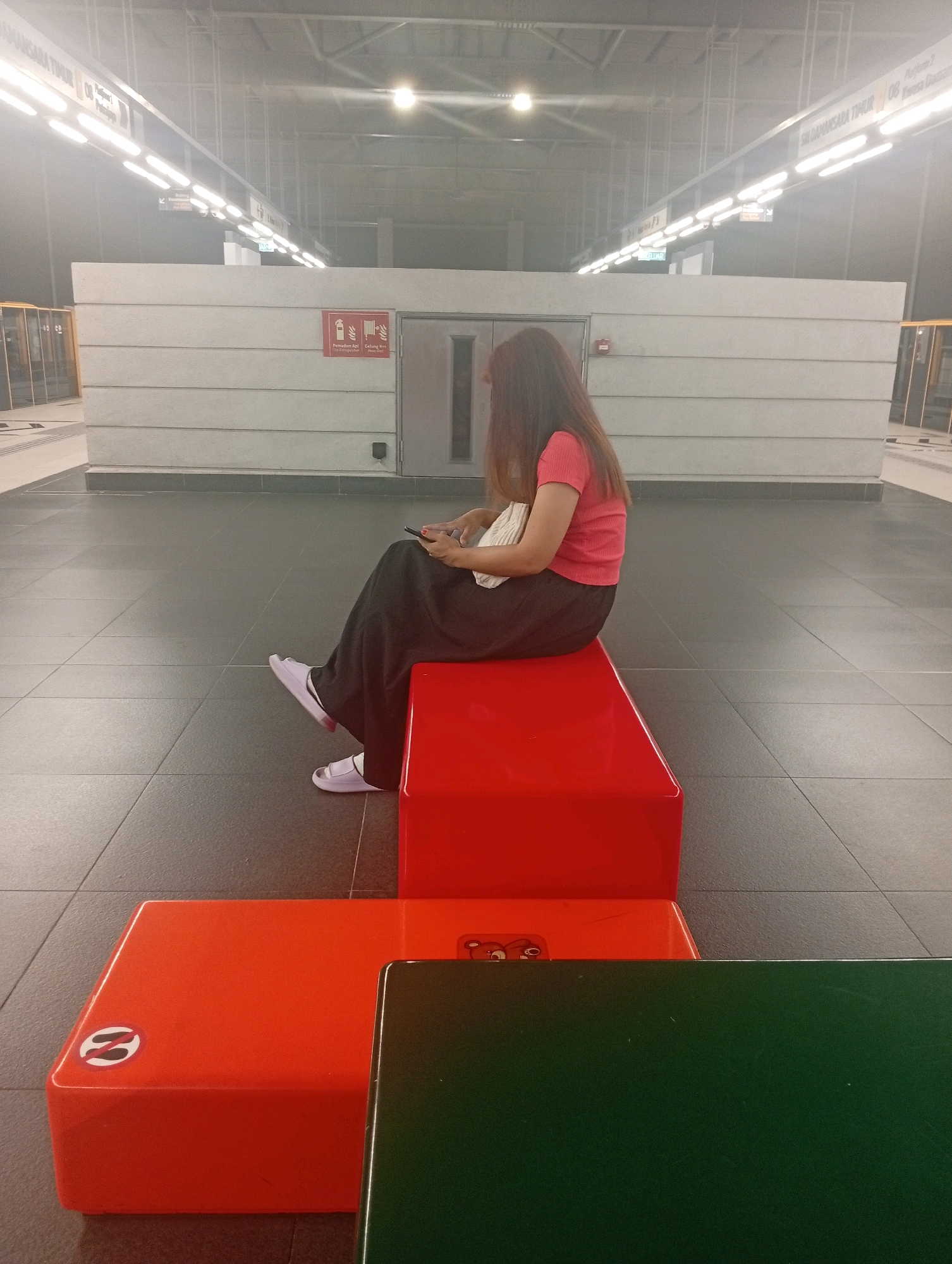 A woman with long brown hair is sitting on a bright red rectangular bench at a train station platform in Petaling Jaya, Malaysia. She is wearing a pink short-sleeved shirt and dark wide-leg pants, and white sandals. The woman is looking down at her phone, which she is holding in her hands. A light-colored bag rests on her lap. The platform is clean and modern, with a tiled floor. Overhead, fluorescent lights illuminate the space, and signs indicate platform numbers and destinations, one reading "SJLANAAN TIMUR." In the background, there is a concrete wall with a fire extinguisher sign and a metal door. In the foreground, there are two other benches, one orange and one dark green. The orange bench has a circular logo with a stylized "n" inside it. The overall atmosphere is one of quiet waiting.