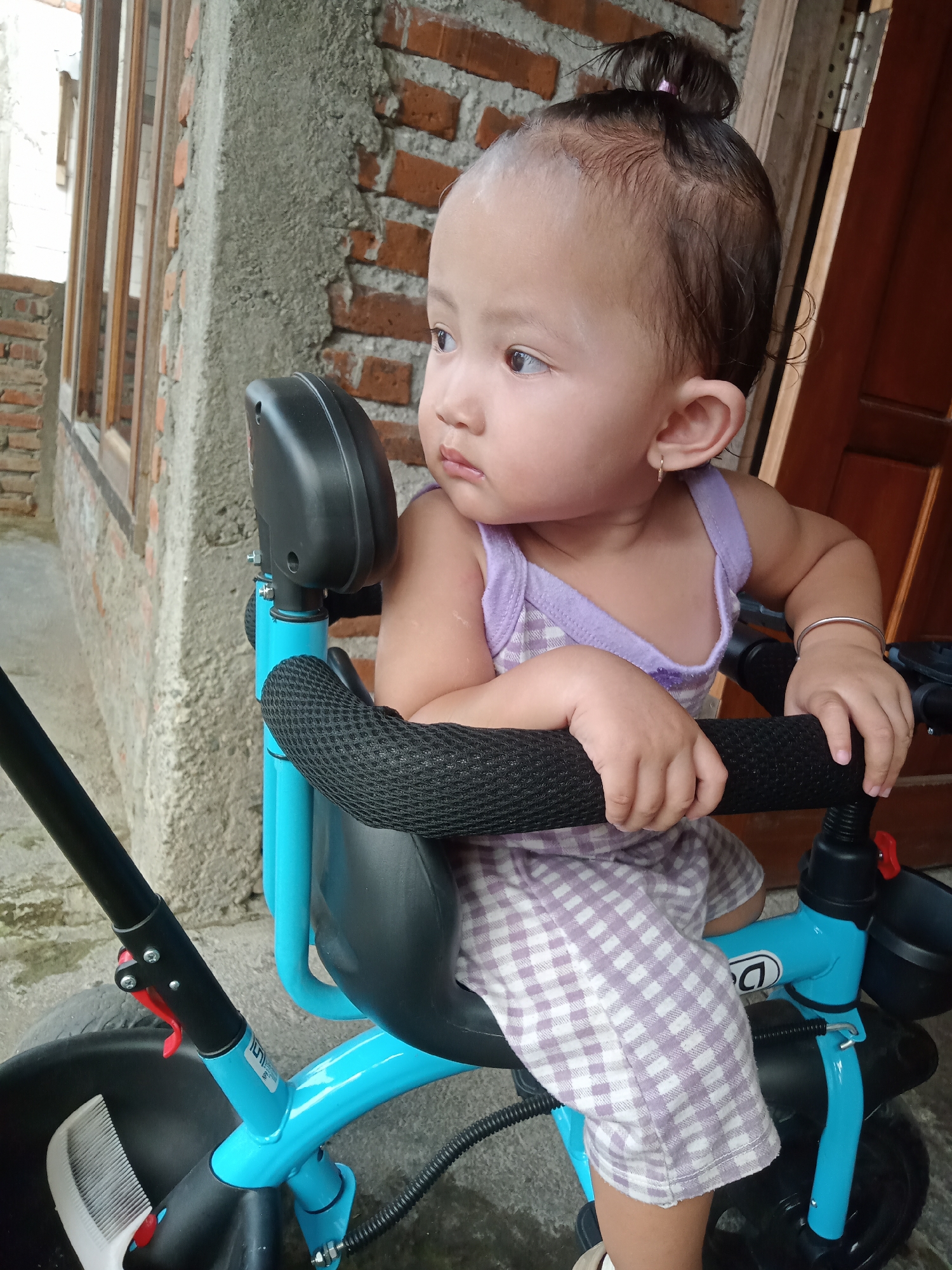 A toddler sits on a bright blue tricycle in Bungur, Indonesia. The child is wearing a lavender and white checkered dress and a lavender tank top. The toddler's hair is pulled up into a small bun. The tricycle has a black padded handlebar and a black backrest. The background shows a brick building and a wooden window frame.