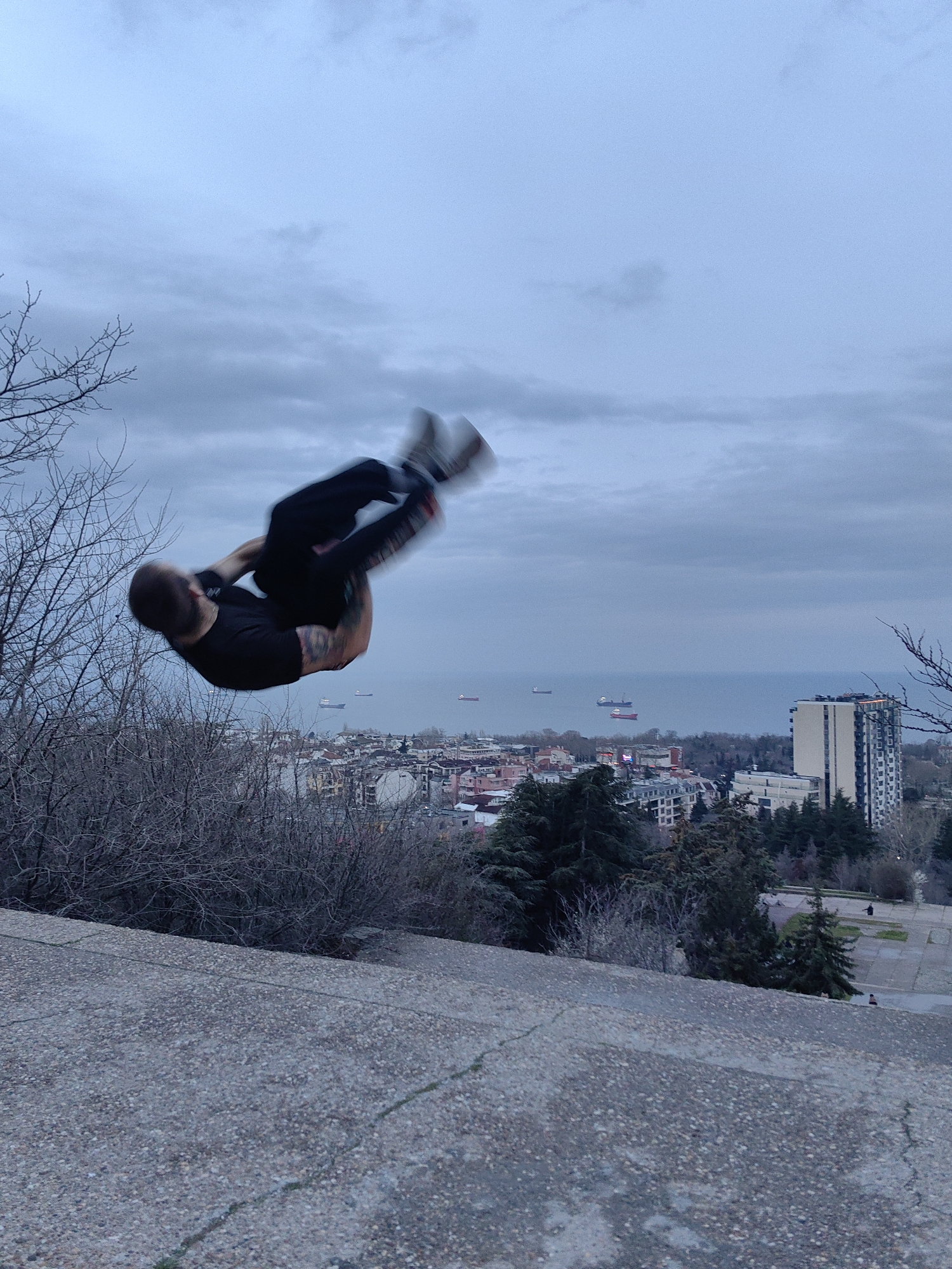 A blurred image captures a person performing a backflip in Varna, Bulgaria. The individual, dressed in a black shirt and dark pants, is mid-air with arms tucked and legs bent, suggesting rapid rotation.
The backdrop includes a cityscape with various buildings, some interspersed with green trees. A tall, pale building stands out. Further back, the sea is visible with several ships in the distance under a cloudy sky.
In the foreground, a gray concrete surface with a pebbled texture angles downward. To the left, skeletal tree branches protrude into the frame. The scene appears to be shot from an elevated position, likely a park or an area overlooking the city and the Black Sea.
