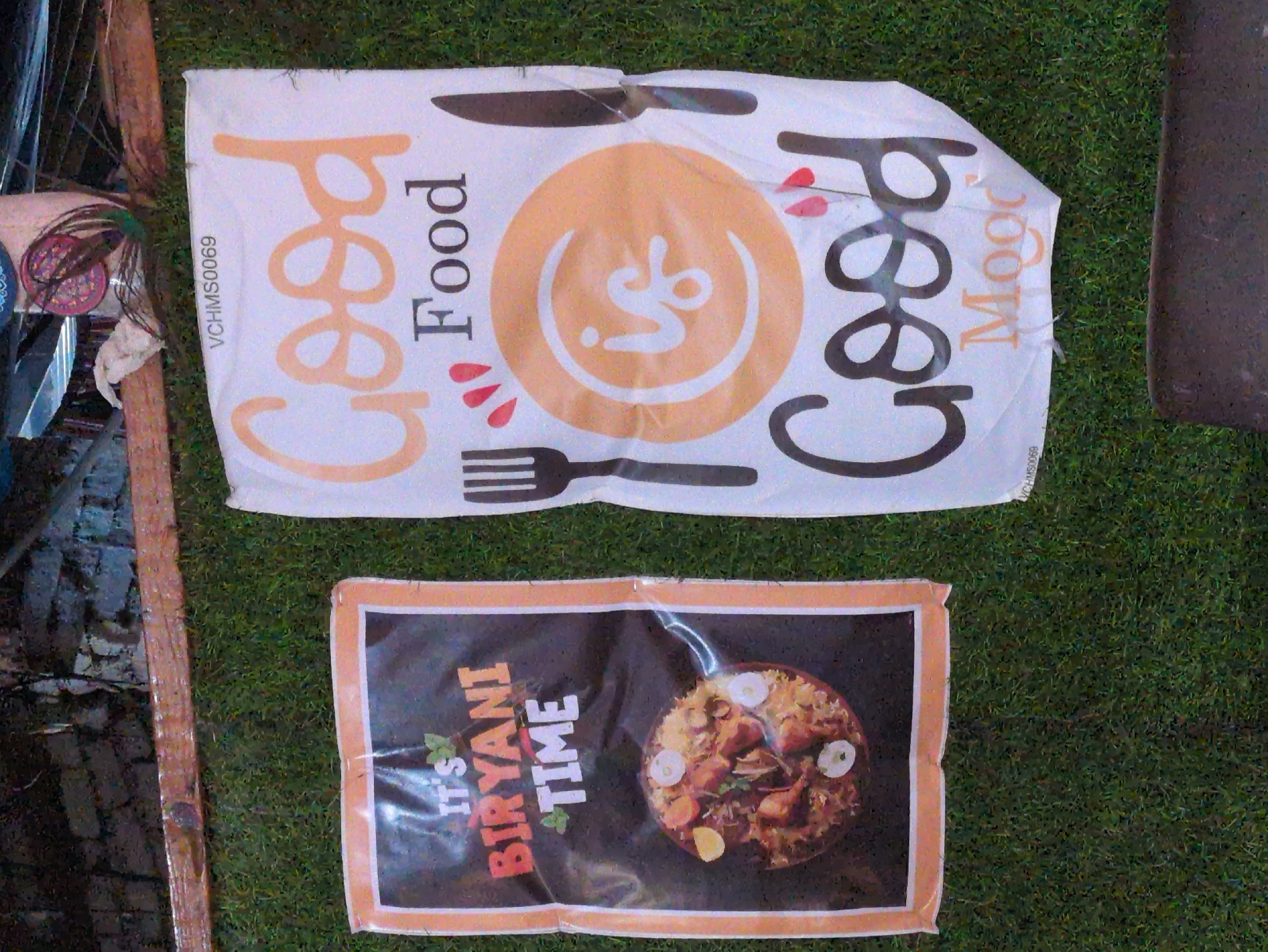 The image depicts two signs displayed outdoors against a backdrop of artificial grass and a stone wall. The sign on the left, framed in orange and white, is dark with the text "IT'S" in white and "BIRAYANI TIME" in orange and white with a leaf motif. Below the text is a circular image of a biryani dish. The sign on the right is larger and predominantly white, featuring the text "Good Food is Good Mood". The word "Good" is written in large, orange, stylized font, followed by "Food" in black. In the center, a circular orange design with a white "is" inside is present, accompanied by a fork and knife graphic on either side. The signs are positioned on a grassy surface, possibly a wall or a display area, with a wooden beam running horizontally above them. The image appears to have been taken in a public space, possibly a street or an outdoor dining area in Bhagsu, India, given the context of the biryani advertisement and the overall informal setting.