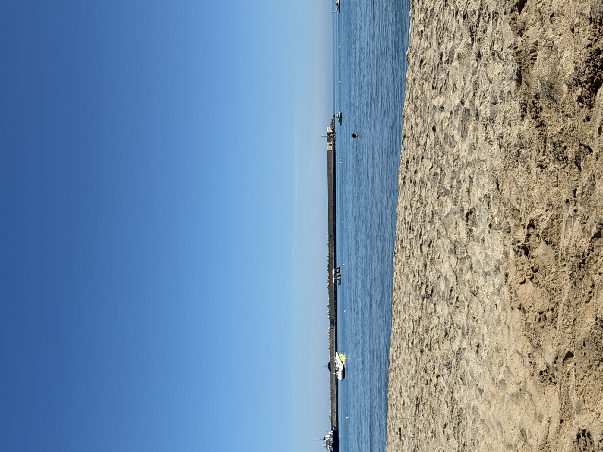 The image shows a beach and the sea on a clear day, likely in the city of Rethymno, Greece, given the context of the provided hints. The foreground is dominated by sand dunes, with intricate patterns of light and shadow. Beyond the sand is the calm, blue sea, stretching towards the horizon. In the distance, a long breakwater or pier extends into the water, with a lighthouse structure visible on its far end. Several boats are scattered across the sea, including a white speedboat with yellow accents, a smaller boat with an awning, and what appear to be jet skis. One person is visible swimming in the water near a jet ski. The sky is a vast expanse of clear, bright blue. The overall scene depicts a peaceful coastal environment.