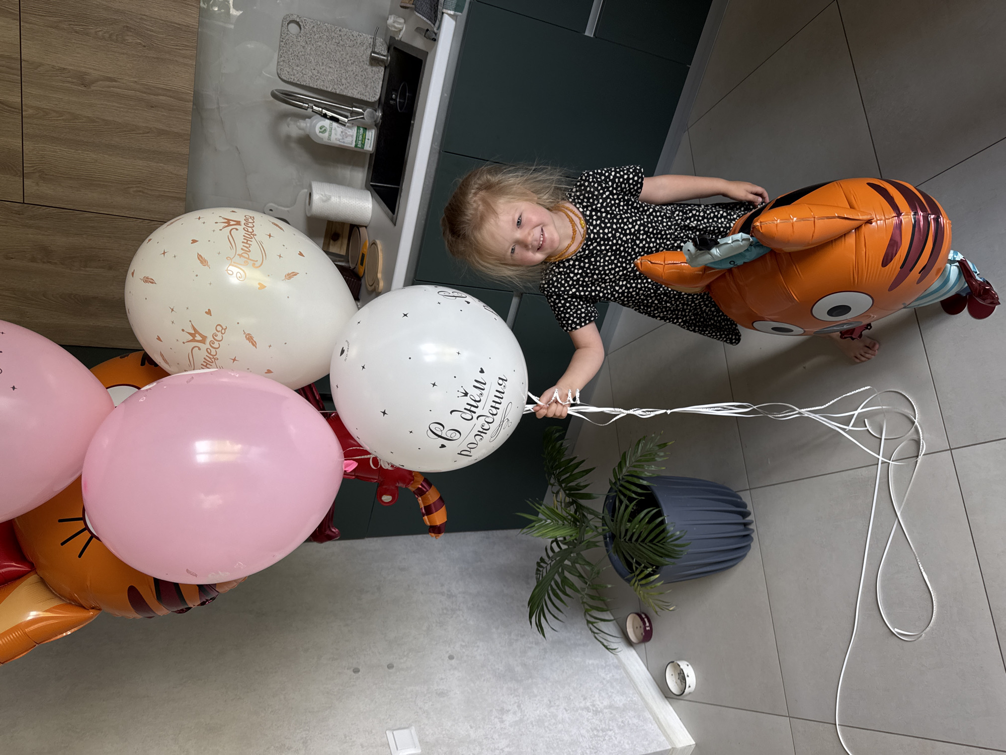 A young girl with blonde hair and fair skin, wearing a black and white polka dot dress and an amber necklace, stands in a modern kitchen, smiling at the camera. She is holding a cluster of balloons, including a large white one with black text and stars, a pink balloon, and an orange foil balloon shaped like a tiger. Behind her, kitchen cabinets with a wood grain finish are visible, along with a sink, faucet, and countertop. To her left, a potted palm plant sits on the tiled floor. The overall lighting suggests an indoor setting, possibly during the day.