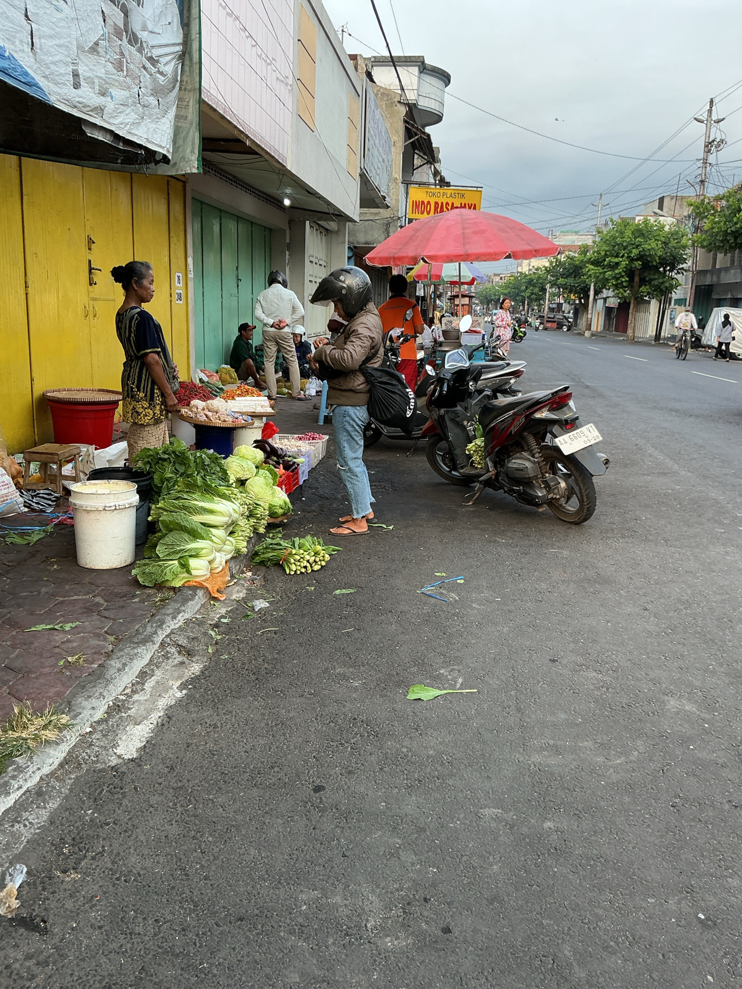 An outdoor street scene in Magelang, Indonesia, depicts a roadside market with vendors and customers. On the left, a woman in a traditional outfit stands near a yellow storefront, overseeing a display of fresh vegetables and other goods. To her right, a person in a brown jacket and jeans with a helmet on their head is standing near a red motorcycle, which is parked on the asphalt road. The road is lined with stalls covered by red umbrellas, selling various items. In the background, other vehicles and people are visible, and buildings line the street. The sky is overcast, suggesting a cloudy day.
