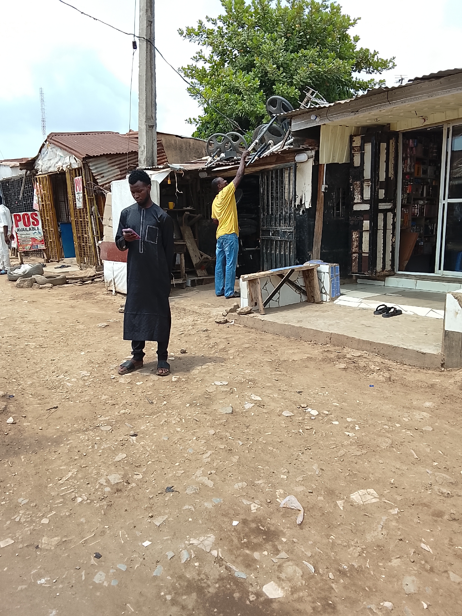 The image shows a dusty, uneven dirt road in what appears to be a commercial area in Abuja, Nigeria. In the foreground, a man in a long black robe and sandals looks at his cell phone. Behind him, another man in a yellow shirt and blue jeans is reaching up towards the roof of a corrugated metal-roofed structure, possibly adjusting some equipment on the roof. To the left, there are other small buildings and a "POS AVAILABLE" sign. A small concrete platform with discarded sandals in front of a shop with shelves inside is seen, along with other debris scattered on the ground. A bench is beside the shop. A utility pole is adjacent to the buildings.