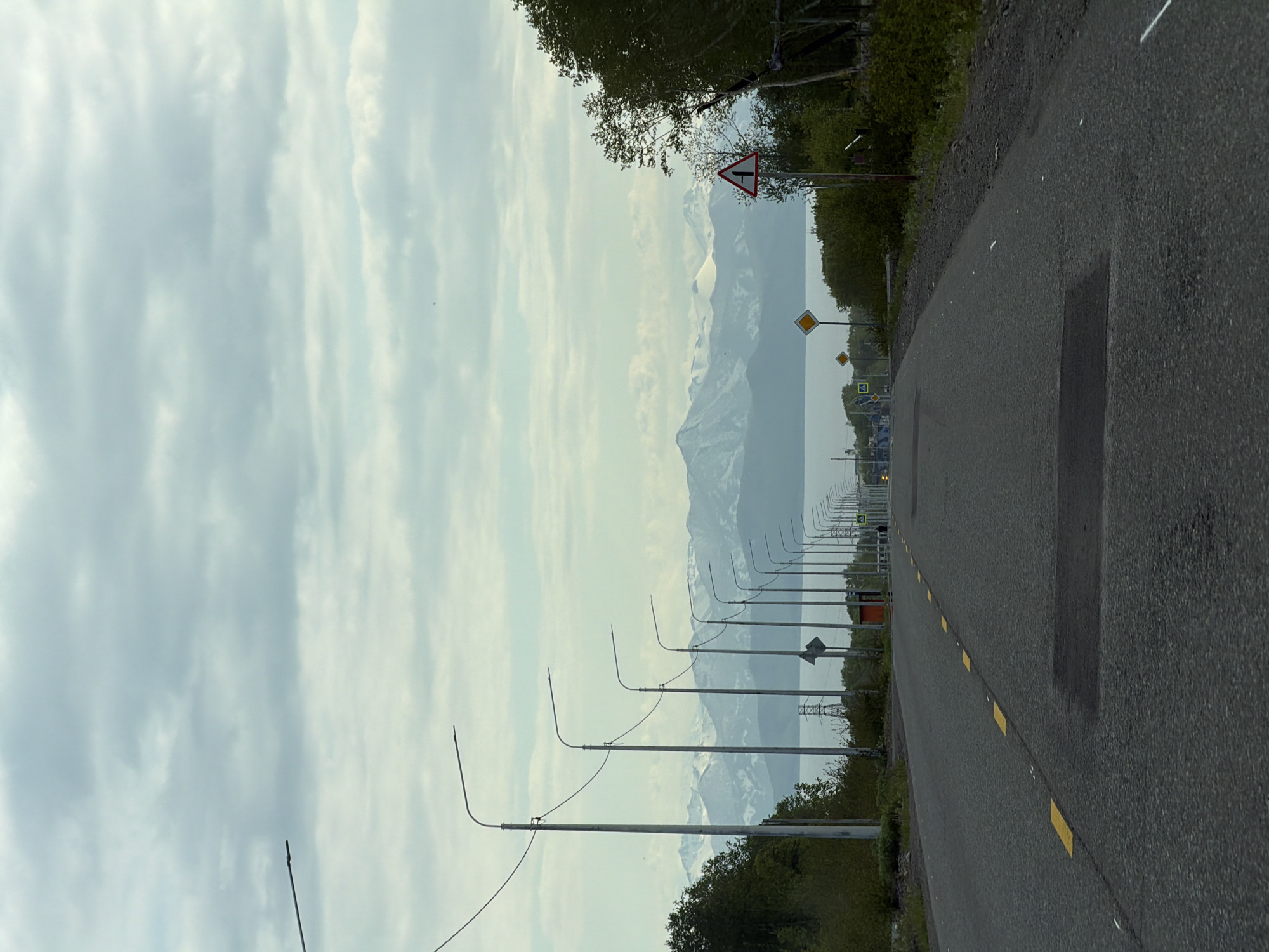 The image depicts an asphalt road with yellow dashed lane markings leading into the distance. The road is lined with numerous lampposts on the left side, and a dense line of trees on the right. In the background, a body of water is visible, beyond which are snow-capped mountains under a cloudy sky. There are no people clearly visible in the scene, though a pedestrian crossing sign is present further down the road on the right. The environment appears to be a semi-urban or rural area outside of the city of Krutoberegovoy, Russia, given the presence of the road, lampposts, and natural landscape. The overall lighting suggests it is daytime, possibly overcast.