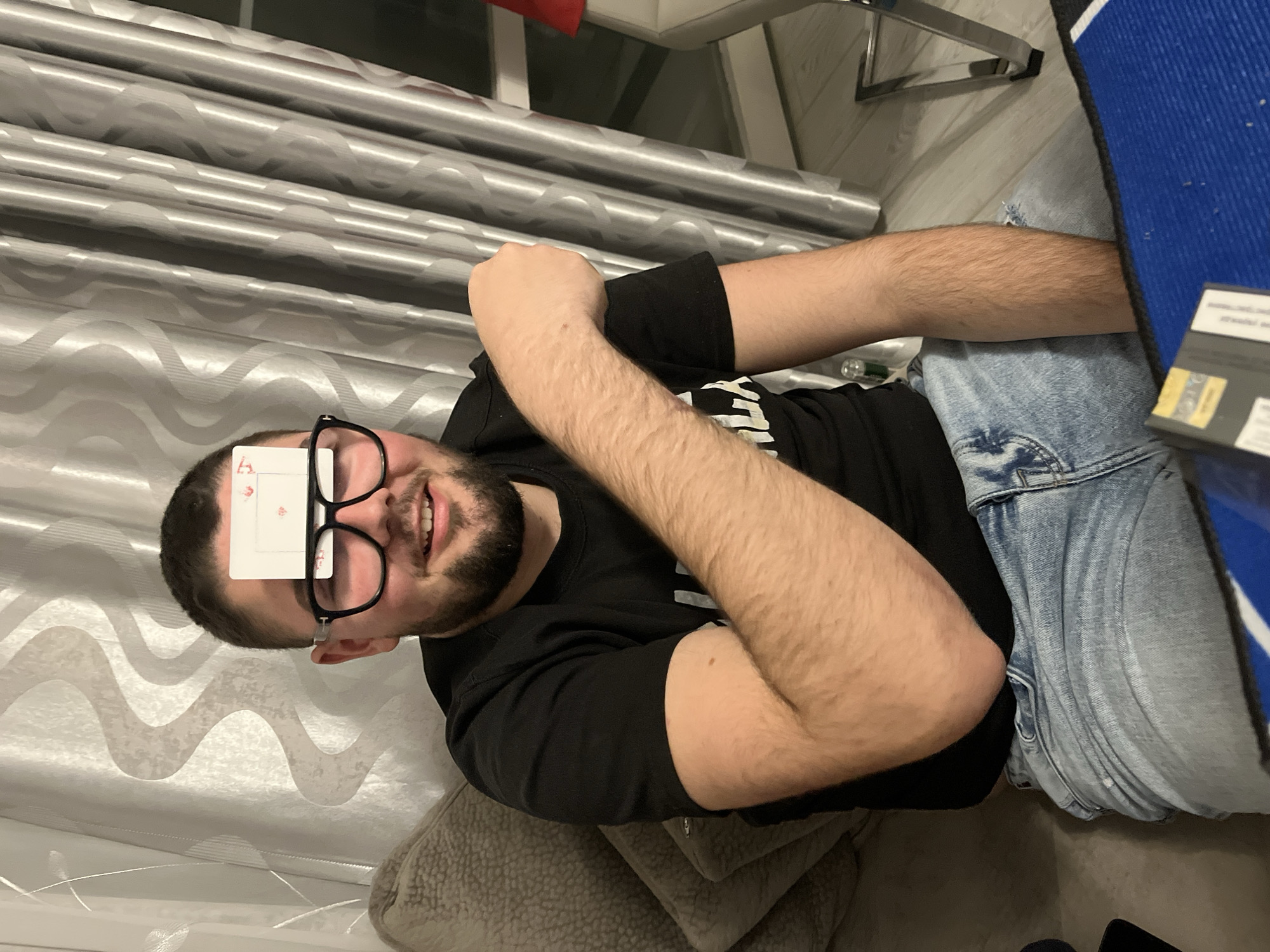 A man with glasses and a beard is lying on his back with a playing card on his forehead. He is wearing a black shirt and blue jeans. He is in a room with a curtain and a chair. The location is not discernible from the image.