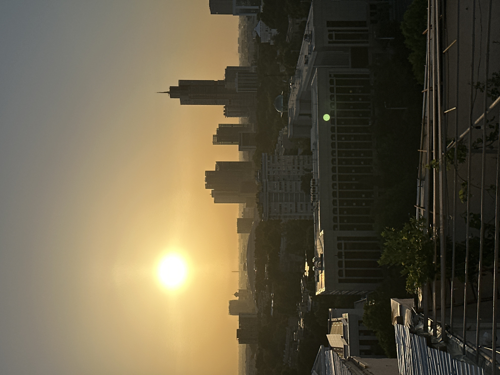 The image depicts a sunset over the city of Tashkent, Uzbekistan, as viewed from a high vantage point, potentially a rooftop or high-rise building. The setting sun dominates the left side of the frame, casting a warm, golden light across the sky and cityscape. The city skyline is composed of various high-rise buildings and smaller structures, partially obscured by the hazy atmosphere. A prominent, tall skyscraper is visible toward the center-left of the image. The foreground includes elements of the building from which the photo was taken, such as a metal railing and portions of the building's exterior. The overall environment suggests an urban setting bathed in the warm glow of the setting sun.