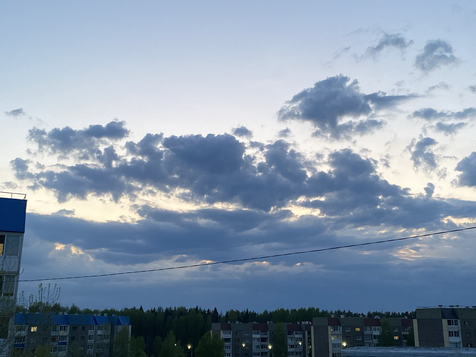 The image depicts a cityscape in Markovskii, Russia, under a cloudy sky at dusk. A series of apartment buildings with lit windows are visible in the foreground, interspersed with green trees. A forest lines the horizon behind the buildings. An electrical wire stretches horizontally across the midground of the image. The sky is filled with gray and white clouds, with glimpses of light breaking through. A blue building is seen in the left side of the image.