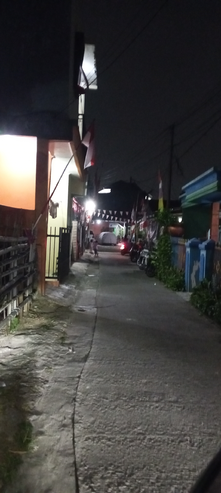 The image depicts a street scene at night in Depok, Indonesia, likely a residential area. A narrow asphalt road runs diagonally from the bottom right to the middle left of the frame. The road is bordered on the left by a low wooden fence and a building with an illuminated orange window. On the right, houses with colorful facades and decorative flags are visible. Several motorcycles are parked along the right side of the road. In the middle ground, a few people are standing near some covered structures, possibly tents. The sky is dark and overcast. The scene is illuminated by artificial lights, creating strong shadows and highlights.
