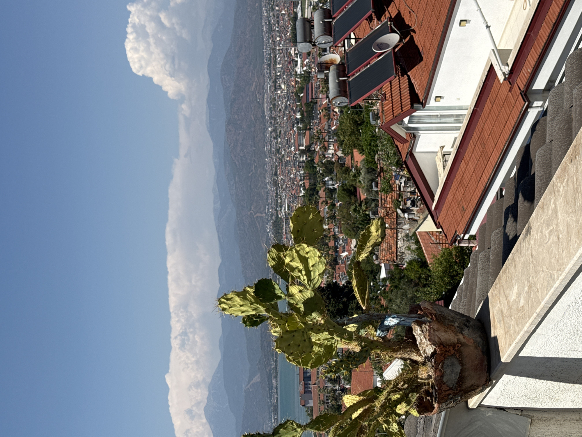 This media file displays a sunlit outdoor scene with a clear blue sky. In the foreground, a prickly pear cactus in a rustic pot is prominently featured. Behind the cactus, a sprawling town with numerous red-tiled roofs stretches towards a body of water and distant, hazy mountains. The buildings appear to be clustered densely together, with some trees interspersed throughout the urban landscape.
On a rooftop to the right, several solar water heaters are visible, suggesting a focus on renewable energy. A white building with a red-tiled roof is also part of the rooftop structure. The scene is bathed in sunlight, casting shadows and highlighting the textures of the cactus and the rooftop elements. The overall impression is one of a warm, sunny day in a coastal or mountainous region. The time of day appears to be late morning or early afternoon, given the angle of the sunlight. There is no visible text in the image. The setting, with its dense housing, red roofs, and coastal mountains, is evocative of a Mediterranean or similar climate, possibly in Türkiye, as suggested by the location context.