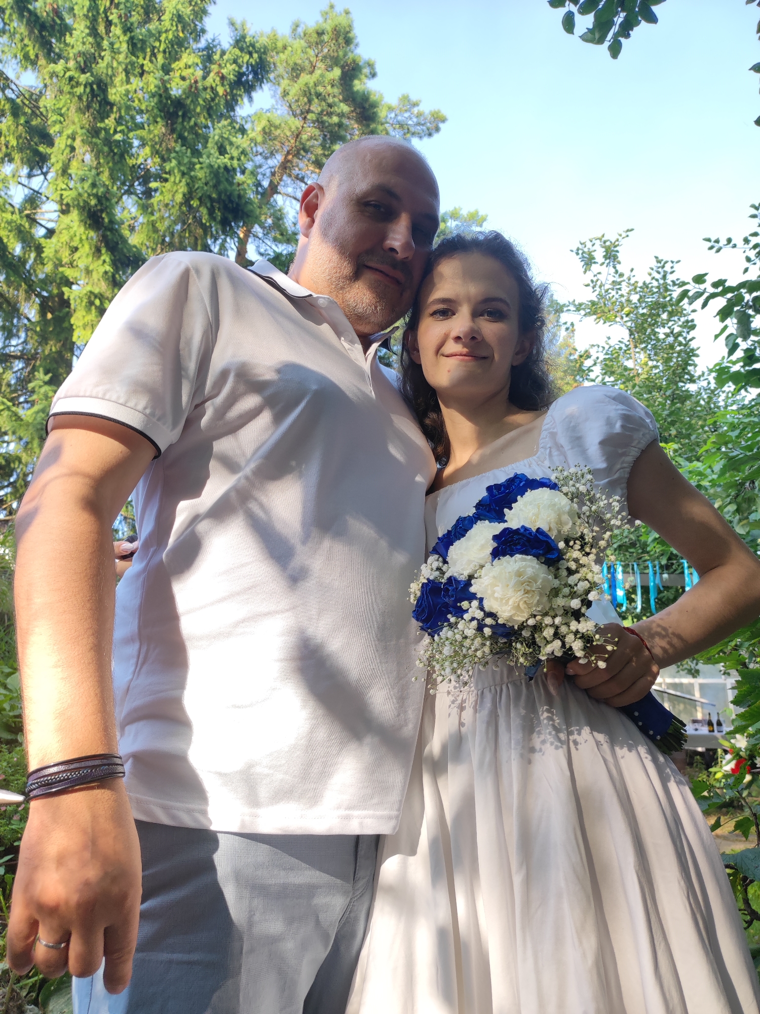 The image shows a couple embracing outdoors. The male is on the left, wearing a white polo shirt and light grey pants, with a bracelet on his left wrist and a ring on his left index finger. The female is on the right, wearing a white dress and holding a bouquet of blue and white flowers. They are positioned in front of trees, with the sky visible in the background. The lighting suggests it is daytime, with sunlight casting shadows. The location appears to be a garden or park setting.