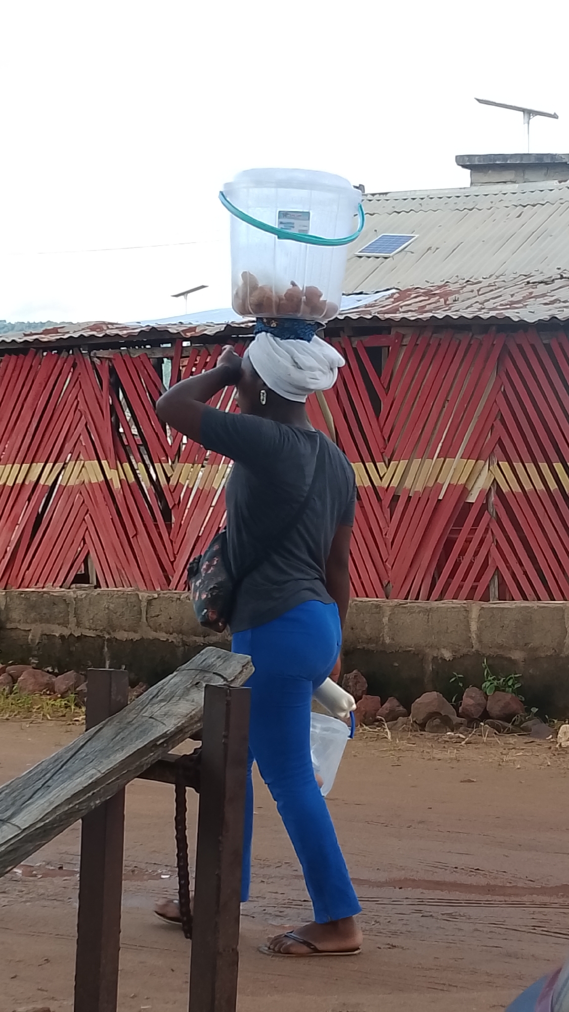 In Adankolo, Nigeria, a woman walks along a dirt road with a large clear plastic bucket containing what appear to be small, brown, round objects balanced on her head. She is wearing a dark gray t-shirt, bright blue leggings, and dark flip-flops. A white cloth is wrapped around her head, supporting the bucket. A patterned bag is slung over her left shoulder. In her right hand, she carries a white plastic bag. Behind her, a building with a corrugated metal roof features a decorative facade of red and yellow diagonal wooden slats. A solar panel is visible on the roof. The ground is rough, with scattered rocks and some sparse vegetation. A wooden structure with metal supports and a chain is in the foreground. The sky is overcast.
