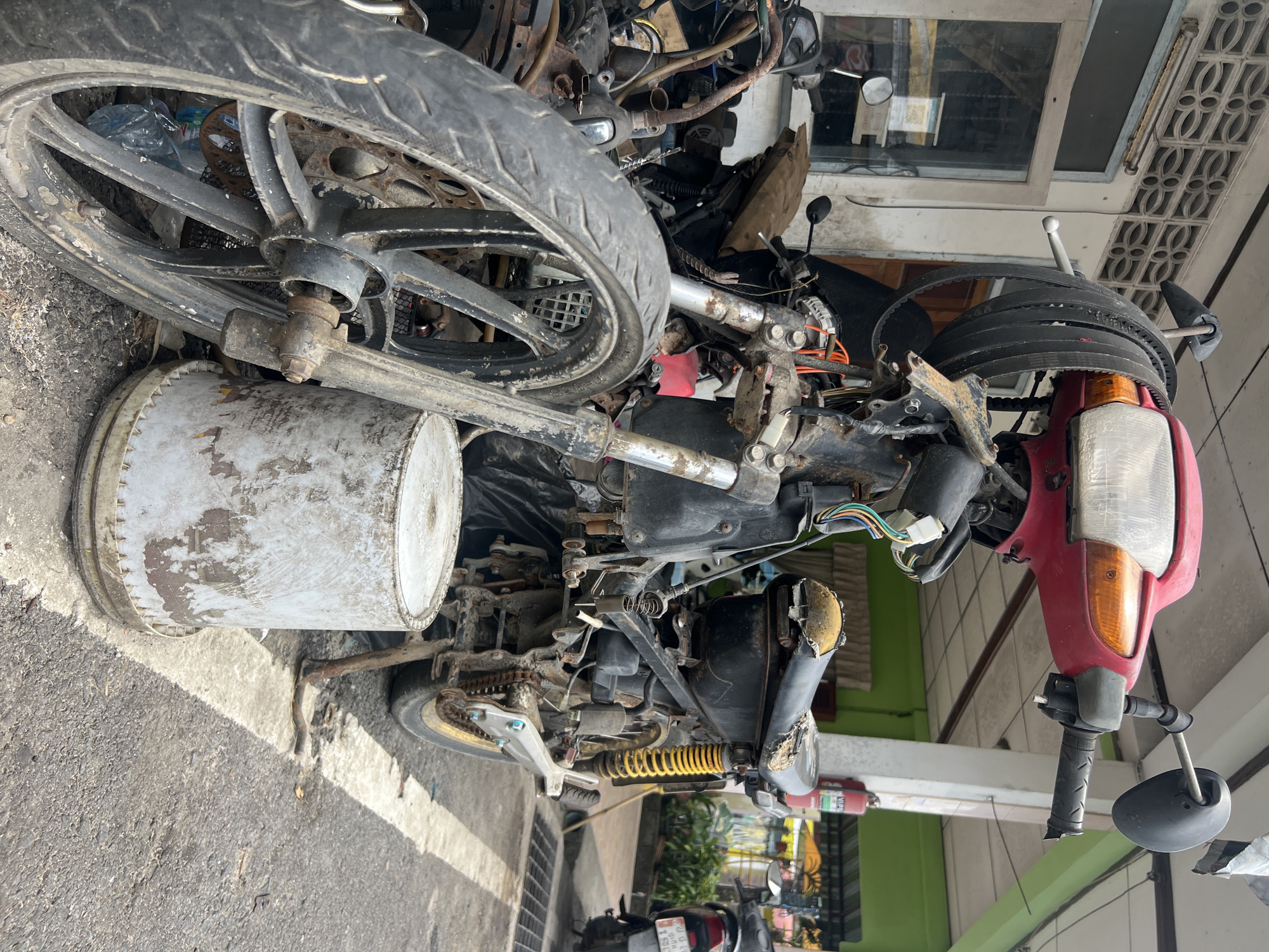 In the city of Choeng Thale, Thailand, a heavily damaged and disassembled red and black motorcycle is parked on a paved surface, likely a street or alleyway, next to a building. The motorcycle appears to be an older model, with its front end partially exposed and various components, including wiring, a drive belt, and the engine, visible. The seat is torn and worn, and the frame exhibits signs of rust and decay. A large, white, cylindrical container, possibly a bucket or drum, stands beside the motorcycle, and other discarded motorcycle parts are scattered around. The building in the background has a tiled roof and a green facade, with a fire extinguisher mounted on a white column. Another motorcycle, less damaged, is parked in the background. The overall scene suggests a repair shop or a storage area for discarded vehicles.