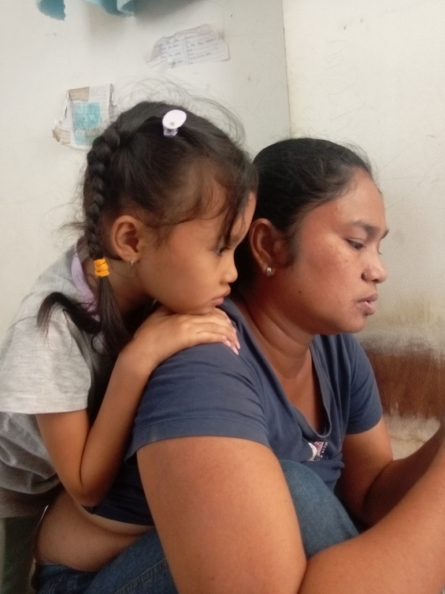 The image depicts a close-up of two females, presumably a mother and daughter, in an indoor setting. The younger female, with braided hair secured by a white clip and yellow bands, leans on the shoulder of the elder female. The elder female is wearing a dark blue t-shirt and jeans. They appear to be indoors, with a plain, light-colored wall in the background. Some papers or posters are affixed to the wall. The lighting suggests it is daytime. The scene is intimate and personal. The background details are minimal, focusing on the interaction between the two individuals. The location is specified as Batam, Indonesia.