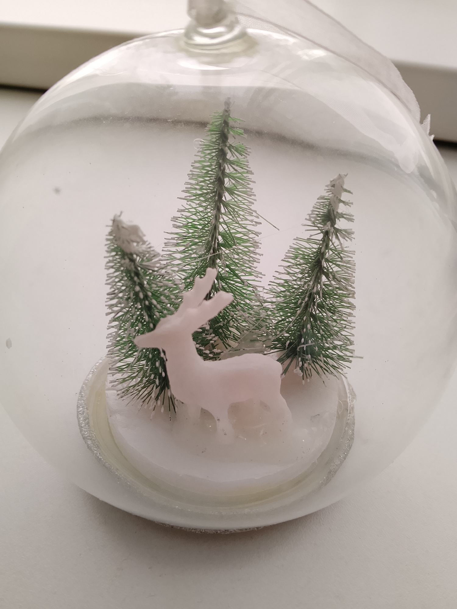 This is a close-up, eye-level, indoor photograph of a clear glass ornament, possibly a Christmas bauble. Inside the ornament, a small winter scene is depicted. Three miniature green bottle brush trees, dusted with white to resemble snow, stand on a white circular base. A pale pink, translucent figurine of a deer stands in front of the trees, facing left and looking downwards. The background is a plain, light-colored surface, likely a table, with a faint light source visible at the top. The ornament itself is suspended by a thin white ribbon, barely visible at the top. No people are present in the image, and no activities are taking place. The image does not provide any information about the city of Abakan, Russia, or its specific location within it.