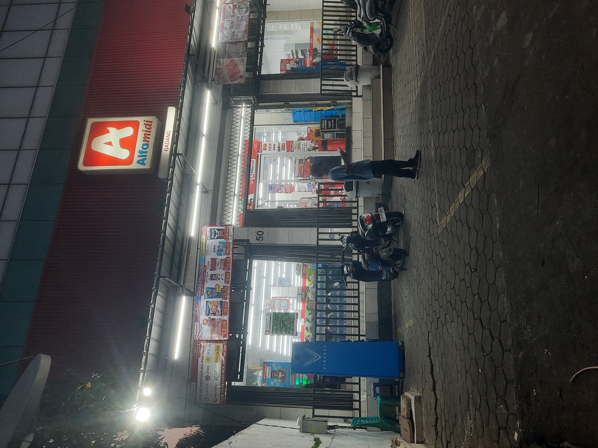 An exterior, eye-level, medium shot shows a Alfamidi convenience store at dusk. The store is located in the Gadang area of North Jakarta, Indonesia. The store's prominent red facade features a large illuminated sign with the Alfamidi logo and name. A smaller sign below the main logo reads "GADANG". The store entrance is open, revealing brightly lit interior aisles stocked with goods. A person stands in front of the store, facing a smartphone, illuminated by the store's lights. Several motorcycles are parked in front of the store, on a paved area. A blue "VINFAST" electric vehicle charging station is visible to the left of the entrance. The ground is paved with interlocking hexagonal stones. The scene is dimly lit, characteristic of evening hours.