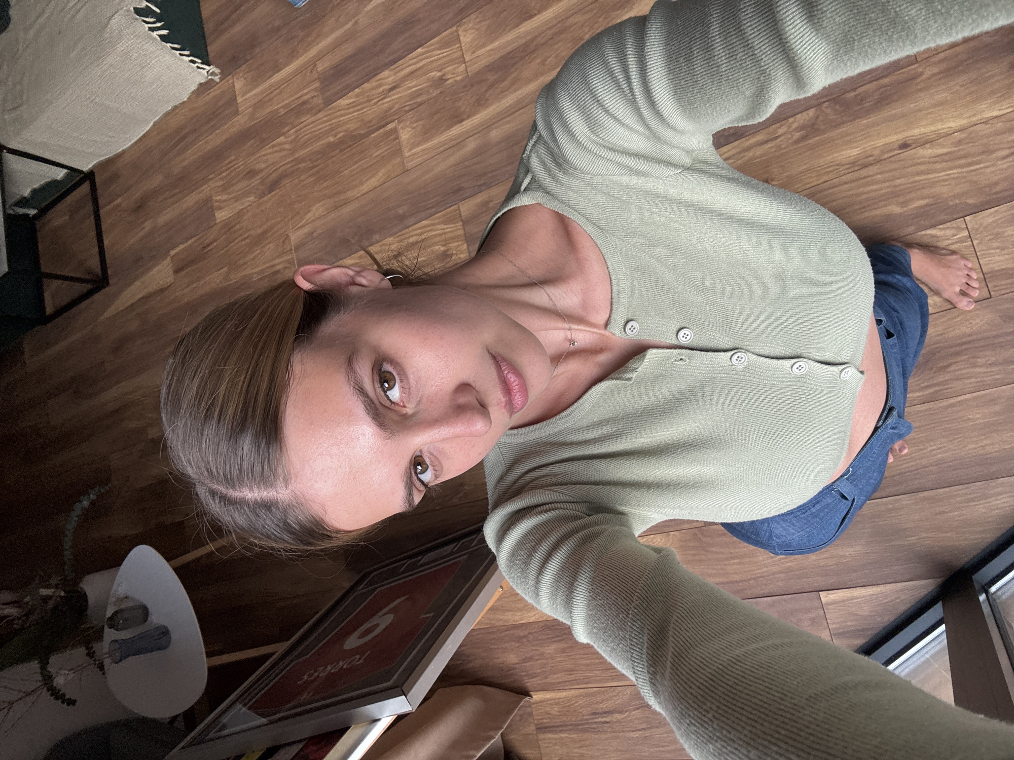 A high-angle selfie captures a young woman looking up directly at the camera, standing indoors on a wooden floor. She has light skin, brown hair styled down her back, and natural-looking light brown eyes. She wears a light green, long-sleeved, ribbed cardigan that is partially unbuttoned, revealing a glimpse of her midriff, and blue jeans. A thin silver necklace with a small pink charm adorns her neck. Her bare right foot is visible in the lower right.
The background features warm-toned wooden floorboards throughout. To the upper left, a beige and dark-toned rug with fringe is partially visible, along with the corner of a dark, metal-framed piece of furniture, possibly a table or stand. In the lower left, near her head, a white, abstract-shaped object holds what appear to be small sticks or tools. Next to this, a framed item leans against something, displaying a red background with a large white number "6" and the word "TORRES" beneath it, both appearing reversed due to the photo's orientation. Natural light, likely from a window partially visible on the far right, illuminates the scene, suggesting it is daytime.