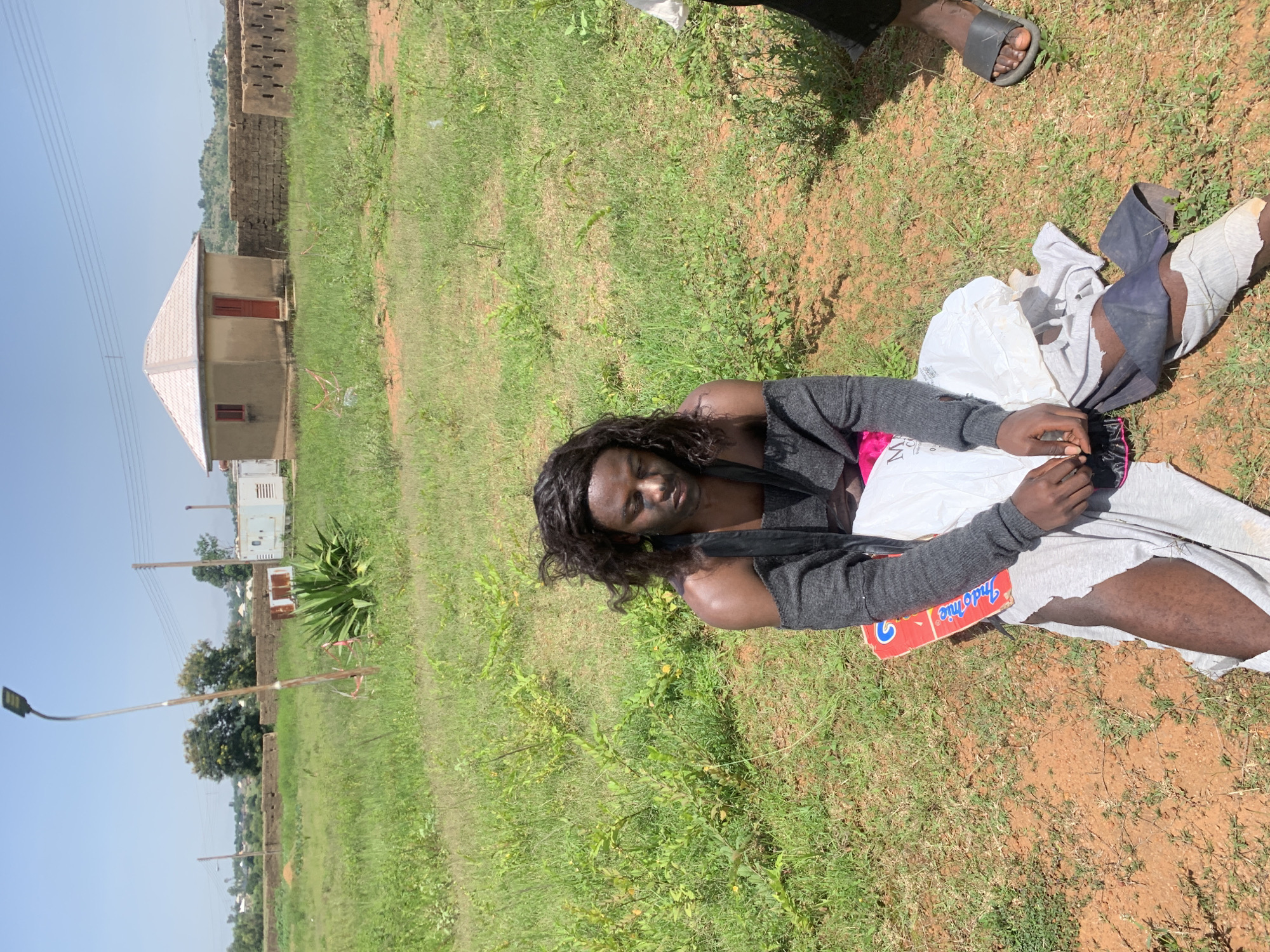 In Gwafan, Nigeria, a person is sitting on the ground in an open, grassy field under a clear sky. The individual, appearing to be male, is wearing a black wig, makeup, a grey off-the-shoulder sweater, and torn white and dark grey pants. The person is seated on what appears to be a red and blue plastic mat. In the background, there is a small, single-story building with a peaked roof and a red door. A brick wall and some scattered vegetation are also visible. A street lamp stands prominently in the left portion of the frame. The overall environment suggests a rural or semi-developed area.