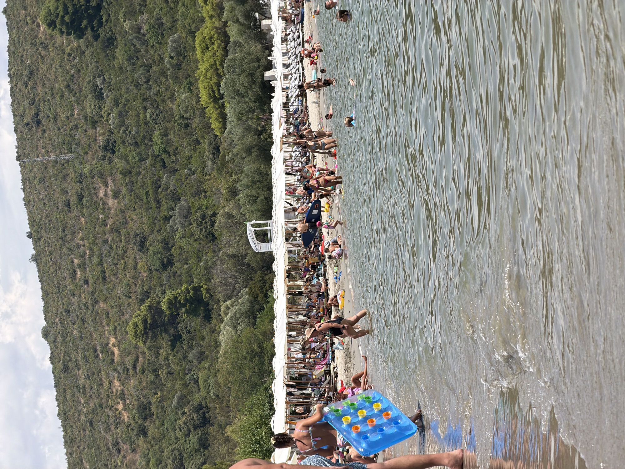 A wide shot shows a crowded beach scene in Bigova, Montenegro, with people sunbathing and swimming. The beach is lined with white umbrellas and lounge chairs, and a lush green hillside rises behind it. In the foreground, several people are wading in the shallow, clear water, with a child in a life vest visible further out. A woman is carrying a blue inflatable raft with orange and yellow circular patterns. The sky is partly cloudy.