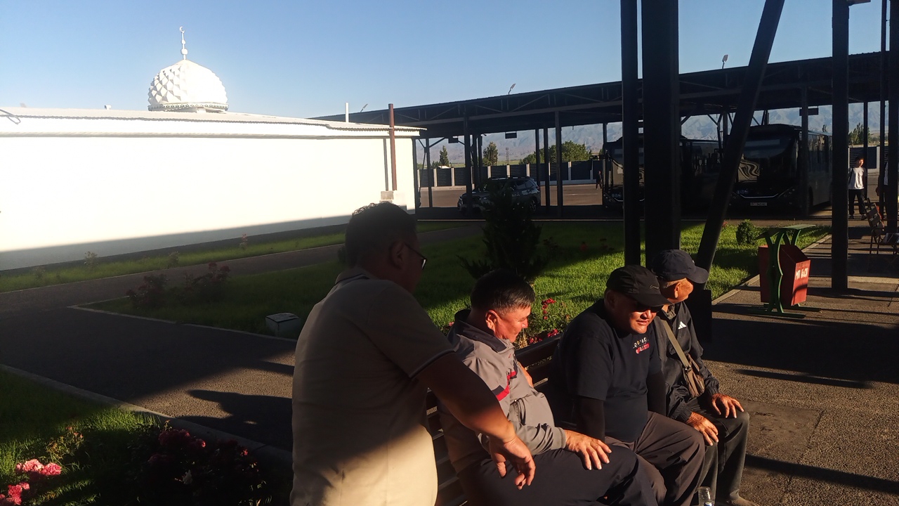 In Balykchy, Kyrgyzstan, four men are seated on a bench outdoors. The man on the far left, wearing glasses and a grey shirt, is facing away from the camera. The second man from the left, wearing a grey jacket, is looking down. The third man from the left, wearing a black cap and black jacket, is looking to his right. The man on the far right, wearing a dark cap and dark jacket, is looking forward. Behind the men, a white building with a dome topped with a crescent moon is visible on the left. A covered structure with support columns, likely a bus shelter or parking area, is in the background, with several dark-colored buses parked beneath it. The ground is paved, and there is grass and some flowering bushes. The sky is clear and blue, indicating daytime.