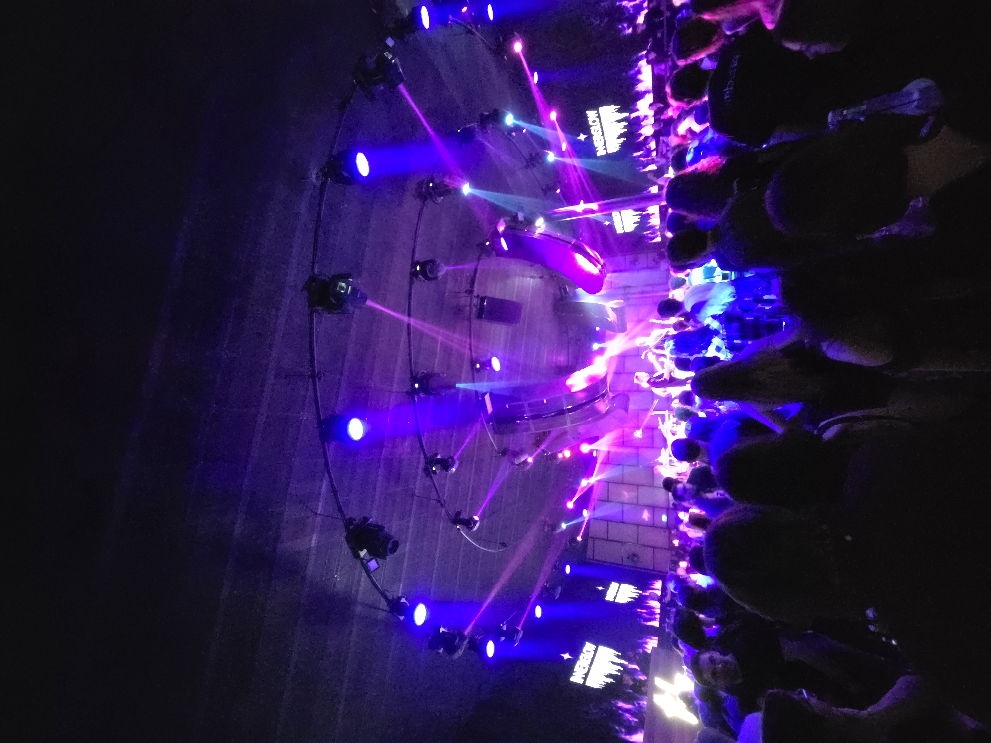 The image captures a nighttime event in what appears to be a tunnel or underground space in Varna, Bulgaria. The tunnel is lit with numerous lights, primarily blue and pink, creating an intense and vibrant atmosphere. A structure with a semi-circular design and evenly-spaced spotlights is a prominent feature. Beams of light project outwards from the structure. Along one side of the image, a crowd of people is visible, their shapes and faces illuminated by the ambient lighting, although details are obscured by the low light conditions. The environment seems to be an urban area based on the architectural characteristics of the tunnel. Text or graphics are faintly discernible on a wall near the crowd.