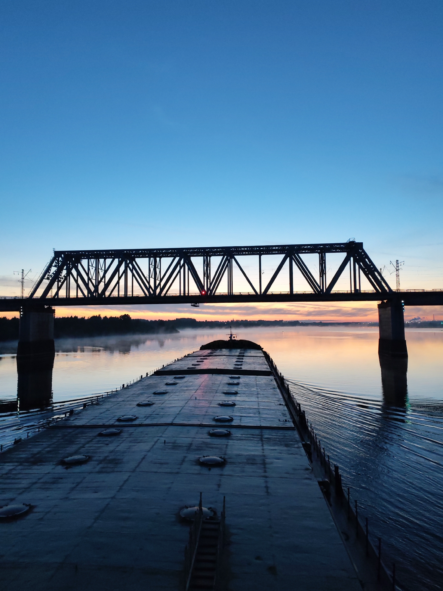 The image shows a large, flat-topped barge navigating a wide river, likely the Volga in the city of Нифантово, Russia, based on the location details. A steel truss bridge spans the river in the background. The scene is bathed in the soft light of dawn or dusk, with a gradient of colors in the sky from blue at the top to orange and pink near the horizon. The river is calm with slight ripples, reflecting the colors of the sky. A light mist hangs over the water near the far shore. The barge occupies the foreground and appears to be heavily laden. The bridge appears to be a railway bridge, with electrical infrastructure near it. The forest line is visible on the horizon between the horizon and the bridge.