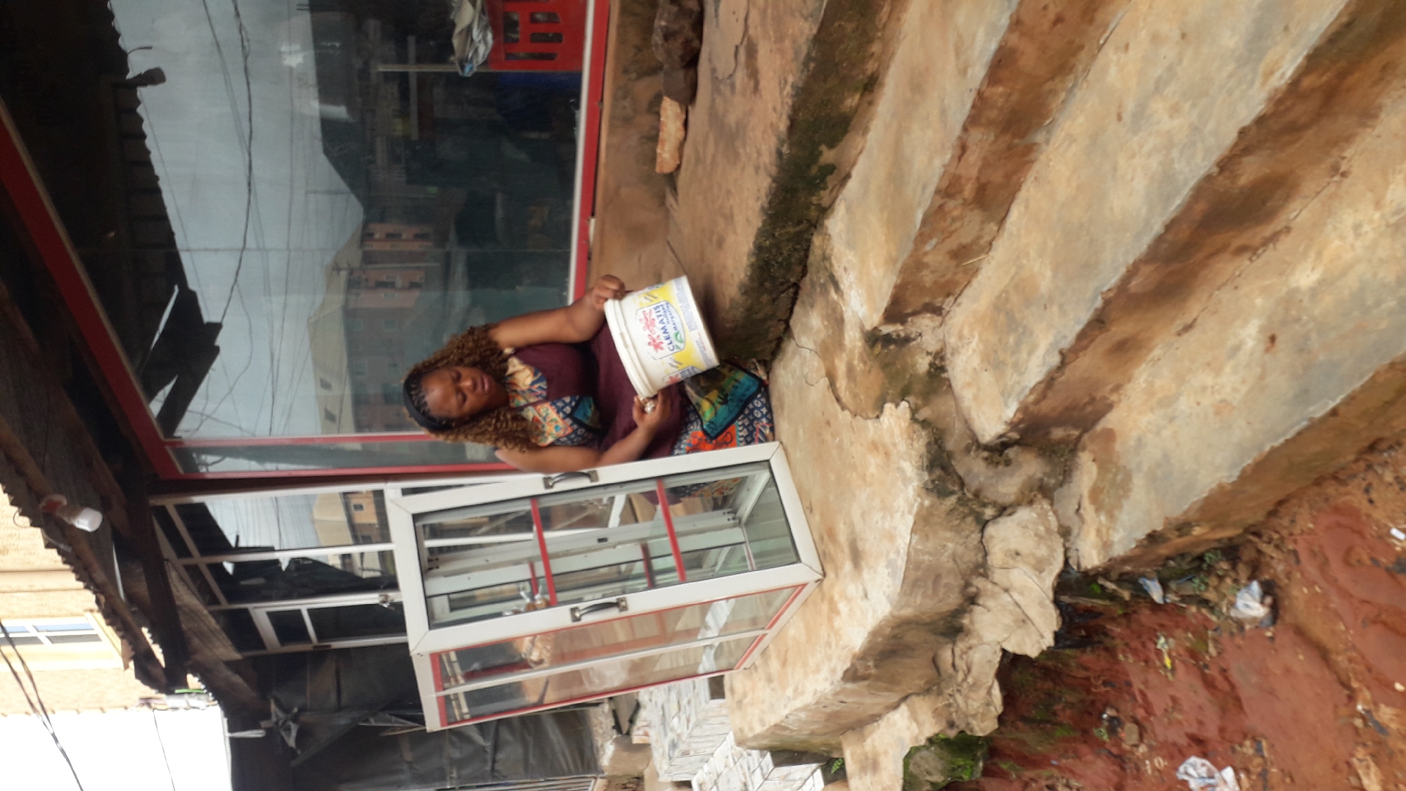 A woman of color, with curly brown hair and wearing a patterned dress, sits on concrete steps in front of a building. To her left is a clear glass display case with a white and red frame, standing upright. The woman holds a white bucket with red and yellow lettering that reads "CLEMATIS." The building appears to be a storefront with large glass windows. The ground around the steps is uneven and covered in red soil, with some litter scattered about. In the background, other buildings are visible through the storefront windows. The scene suggests an outdoor setting, possibly in a commercial area of Nkpor, Nigeria.
