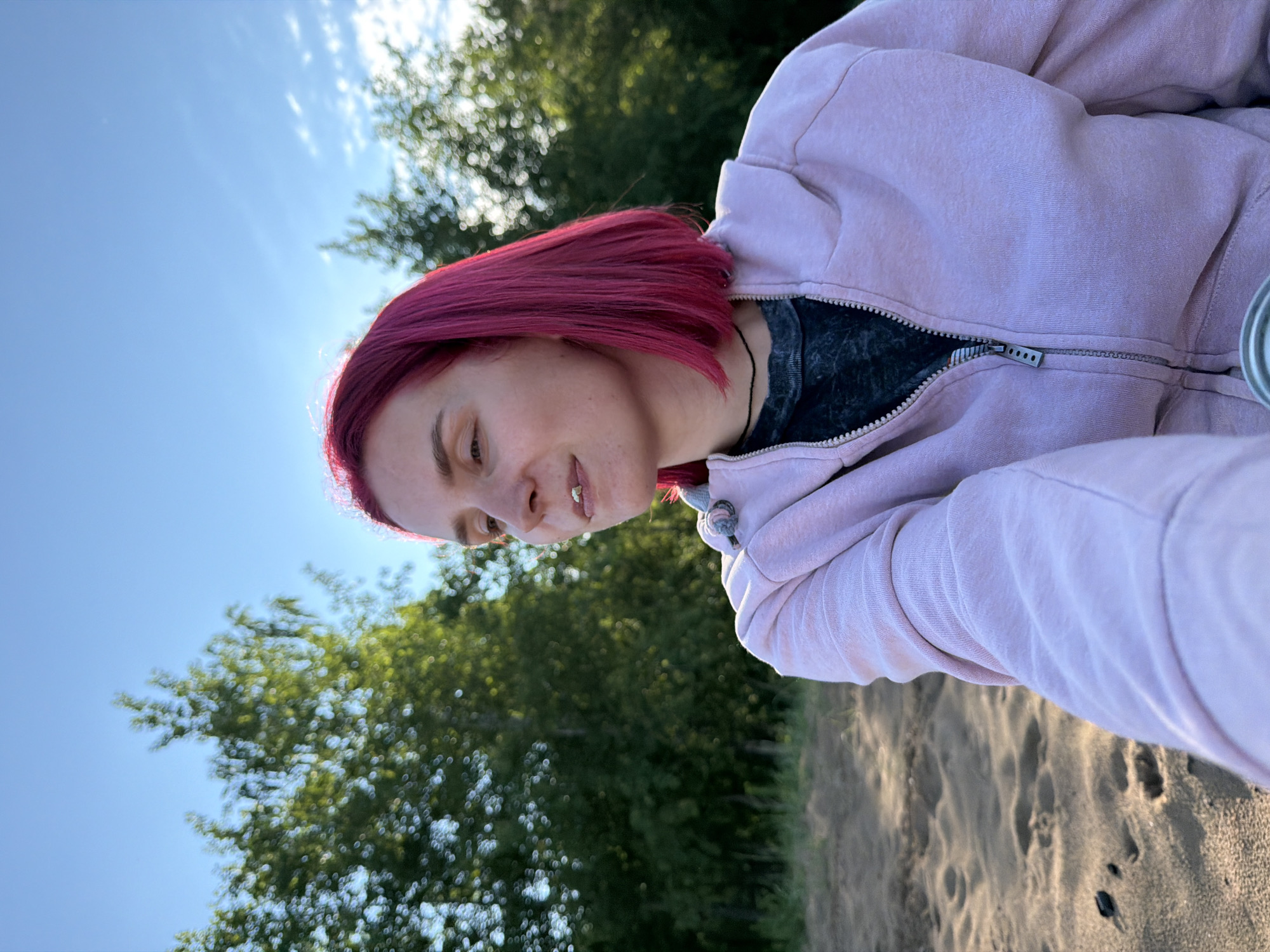 The image, originally rotated 90 degrees clockwise, depicts a young person outdoors under a clear, bright blue sky in Noviy, Russia. The individual, likely female, has short, straight, vibrant magenta-pink hair and is seen in a three-quarters profile, looking downwards and slightly to the left. She wears a light pink or lavender hooded sweatshirt with a silver zipper pull, over a dark grey or black textured t-shirt. A thin black necklace is visible around her neck. Her expression is calm and slightly pensive, with a subtle, gentle smile.
The setting is a natural outdoor environment, possibly a beach or riverside. In the background, the sun is visible behind and slightly to the left of the subject's head, creating a bright halo effect around her hair and a lens flare. Lush green trees occupy the mid-ground on the left side of the frame. The lower left corner and bottom edge show sandy ground with visible footprints and small debris, further indicating an open, natural landscape.
The low angle of the sun suggests the photograph was taken during either early morning or late afternoon, often referred to as the "golden hour," providing soft, warm illumination. The weather appears sunny and clear. No visible text is present in the image. The overall mood conveyed is one of peaceful contemplation in a serene outdoor setting.