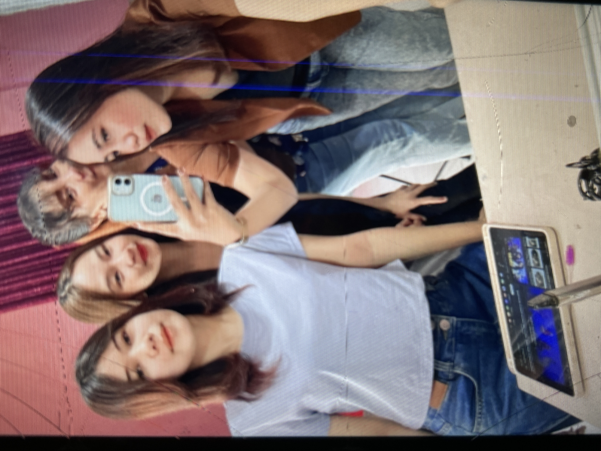 Four young women are posing for a selfie in what appears to be a room with a pink wall and curtains in the background. The woman in the foreground on the left is wearing a light blue t-shirt and jeans. The woman next to her, slightly behind and to the right, has long brown hair and is wearing a brown jacket over a dark top and jeans. Another woman is visible behind them, holding a mobile phone to take the picture, and a fourth woman is mostly obscured behind the others. A tablet is visible on a surface in front of the woman in the foreground, displaying images. The image quality is poor, with visible horizontal lines across it, suggesting it may be a low-resolution scan or a photograph of a screen. The location in Depok, Indonesia cannot be determined from the visual information provided.