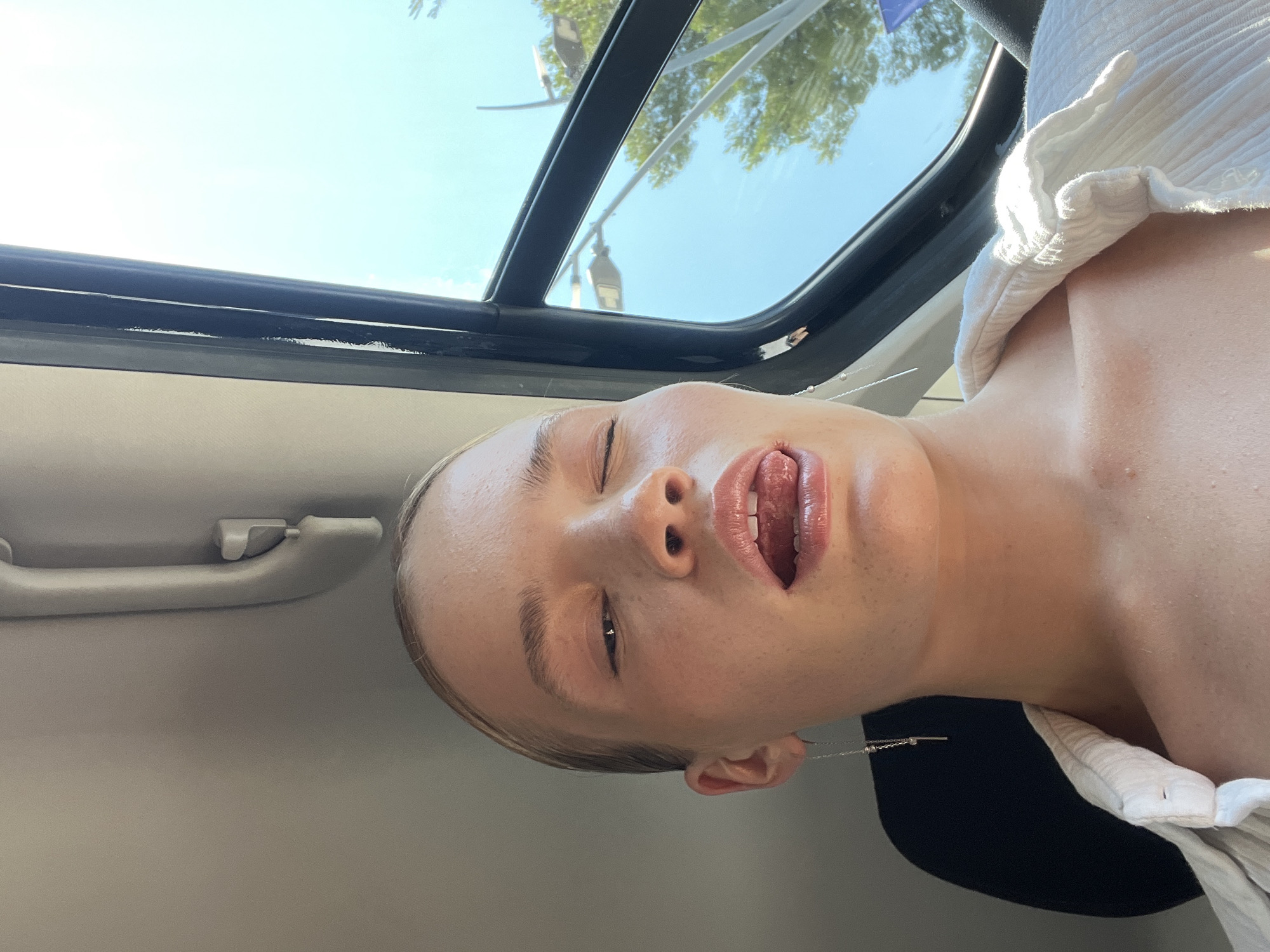 This is a close-up, low-angle, selfie-style photograph taken from inside a vehicle, with the camera oriented sideways. The primary subject is a young person, seen from the nose down, with their head tilted back and to the side. Their eyes are closed, and their mouth is open with their tongue slightly visible. They are wearing a white, textured garment that appears to be a shirt or blouse, and a delicate silver necklace is visible against their neck and collarbone.
The background suggests a sunny day with a bright blue sky visible through a sunroof or open window of the vehicle. Trees and the outline of streetlights can be seen through the glass, indicating an outdoor setting. The interior of the vehicle is visible, with a gray handle on the door or ceiling. The lighting suggests it is daytime, possibly late morning or early afternoon, given the brightness and clear sky. The overall impression is a candid moment, possibly during a car ride, with the subject enjoying the sunlight. No text is visible in the image. The location context provided is Budva, Montenegro.