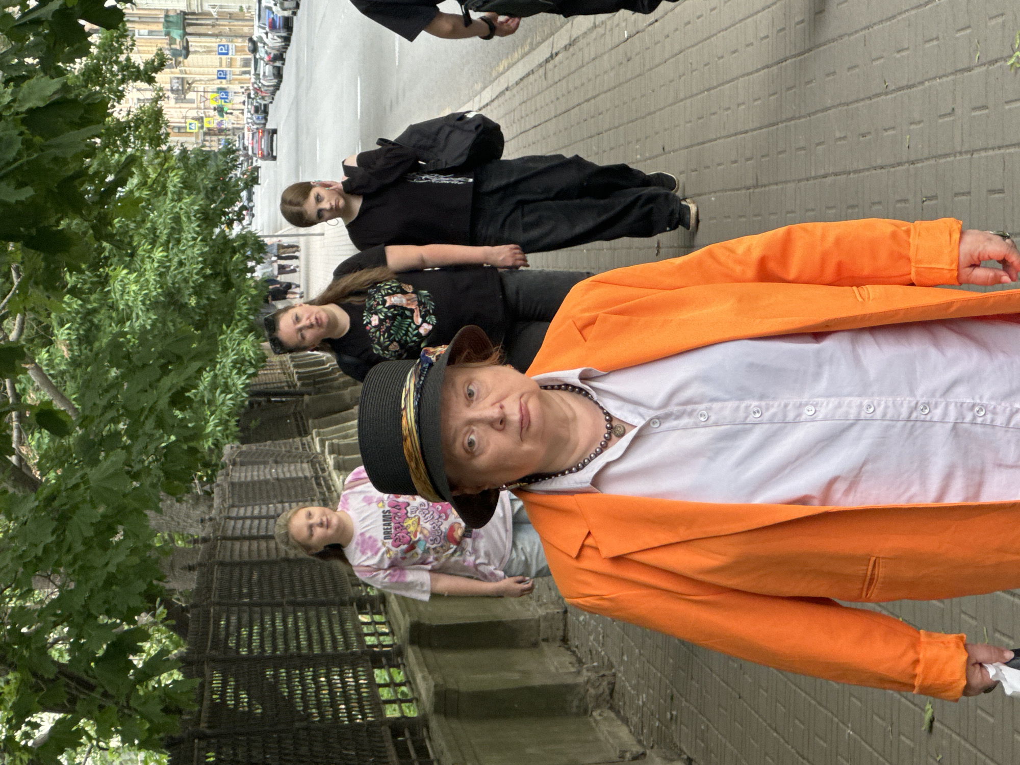 An outdoor, eye-level, full shot captures four people walking on a sidewalk in Saint Petersburg, Russia. The foreground is dominated by a woman wearing a black hat and a bright orange blazer over a white shirt. Her expression is serious as she looks towards the camera. Behind her, three younger individuals walk away from the camera. To her left, a girl in a pink t-shirt with a graphic print and light-colored pants walks. To her right, a woman in a black t-shirt with a floral print and black pants walks. Further to the right, another young woman in a black t-shirt and black pants walks with a black backpack slung over her shoulder, her hand touching her ear. The sidewalk is paved with grey rectangular tiles. A metal fence with a decorative pattern runs along the left side of the sidewalk, separating it from a green, leafy area. In the background, a street with parked cars and buildings can be seen, suggesting an urban environment. The architecture of the buildings indicates a historical European city.