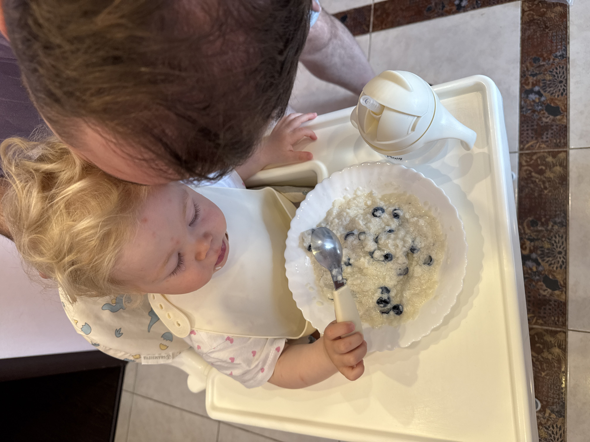 This overhead image depicts an intimate moment between an adult and a young child during a meal in an indoor setting, likely a home in Vladimir, Russia. A man with dark hair, seen from the top of his head and shoulders, leans over a blonde-haired toddler. The child is seated in a white high chair, wearing a cream-colored bib adorned with light blue animal motifs and a white t-shirt with small pink heart patterns.
On the high chair's tray, a white bowl holds what appears to be oatmeal or porridge mixed with blueberries. The child holds a child-sized spoon with a white handle in their right hand, positioned over the food. To the child's right, a white sippy cup is also on the tray. The child's eyes are closed or cast downwards, and their mouth is slightly open. Small red spots are visible on the child's forehead and left cheek. The man's proximity suggests an interaction, possibly feeding the child or offering comfort.
The floor surrounding the high chair is tiled with decorative brown and beige patterns. Visible text includes "SHAMSIYA" on the bib and "РОМИ" (ROMI) on the sippy cup. The scene conveys a quiet, domestic atmosphere during mealtime.