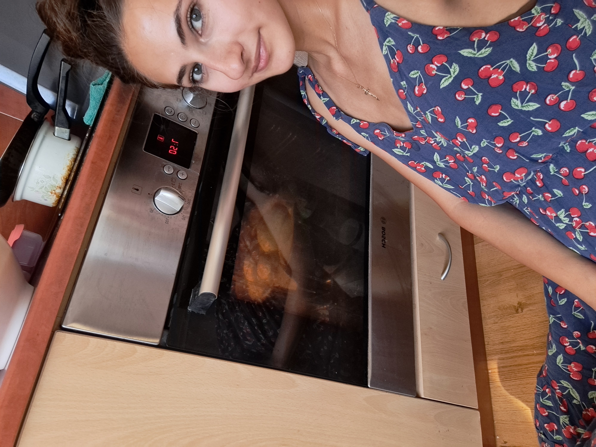 In this close-up, overhead shot, a young woman is seen from the chest up, with her face tilted towards the camera, offering a playful, almost mischievous smile. She is wearing a dark blue dress adorned with a pattern of red cherries. Her gaze is directed at the viewer, and she has captivating grey eyes and dark hair styled up.
The setting appears to be a kitchen, with a stainless steel oven prominently displayed. The oven's digital display shows "02:11". Inside the oven, a golden-brown roasted chicken is visible, suggesting that a meal is in the process of being cooked. The kitchen counter to the left of the oven has a white pot with a rustic pattern, showing some signs of use. The overall ambiance suggests a domestic, home setting. The lighting is natural and diffused, hinting at it possibly being daytime. The woman's expression is relaxed and happy, implying she's enjoying herself or is pleased with the cooking process. The image is oriented such that the woman's head is on the left side of the frame, and her body extends towards the right. A logo that appears to be "BOSCH" is visible on the side of the oven.