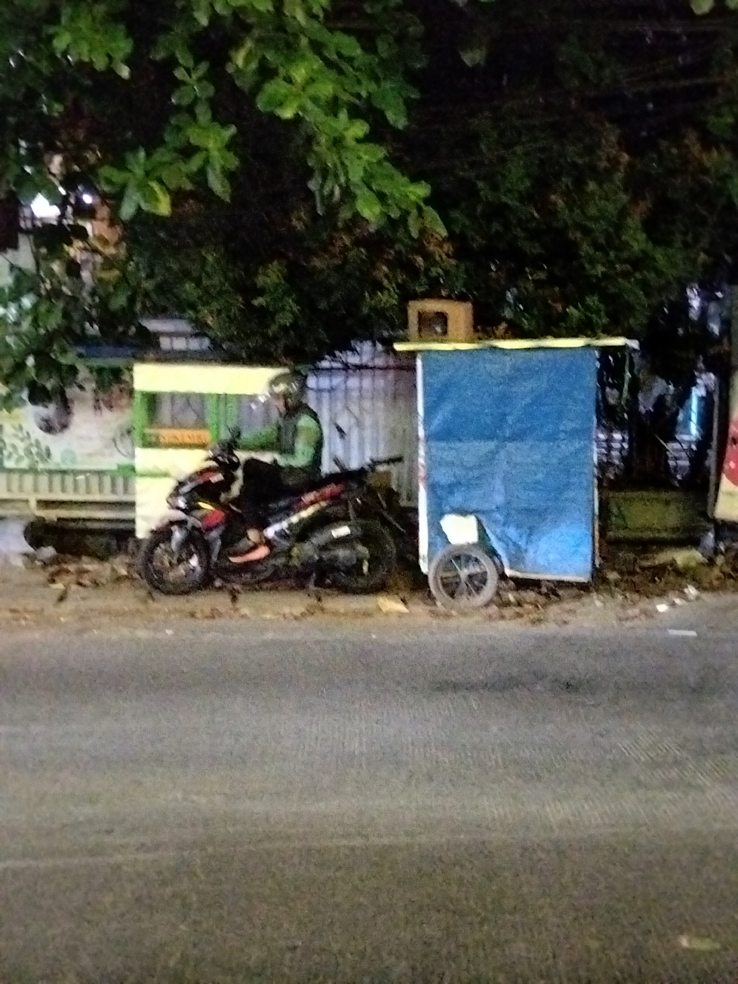 A person wearing a green helmet and jacket sits on a black and red motorcycle. The motorcycle is parked on the side of a road in South Tangerang, Indonesia, in front of a small, makeshift stall covered with blue tarp. The stall has a yellow trim and a small window. Behind the stall are trees. The road surface is asphalt.