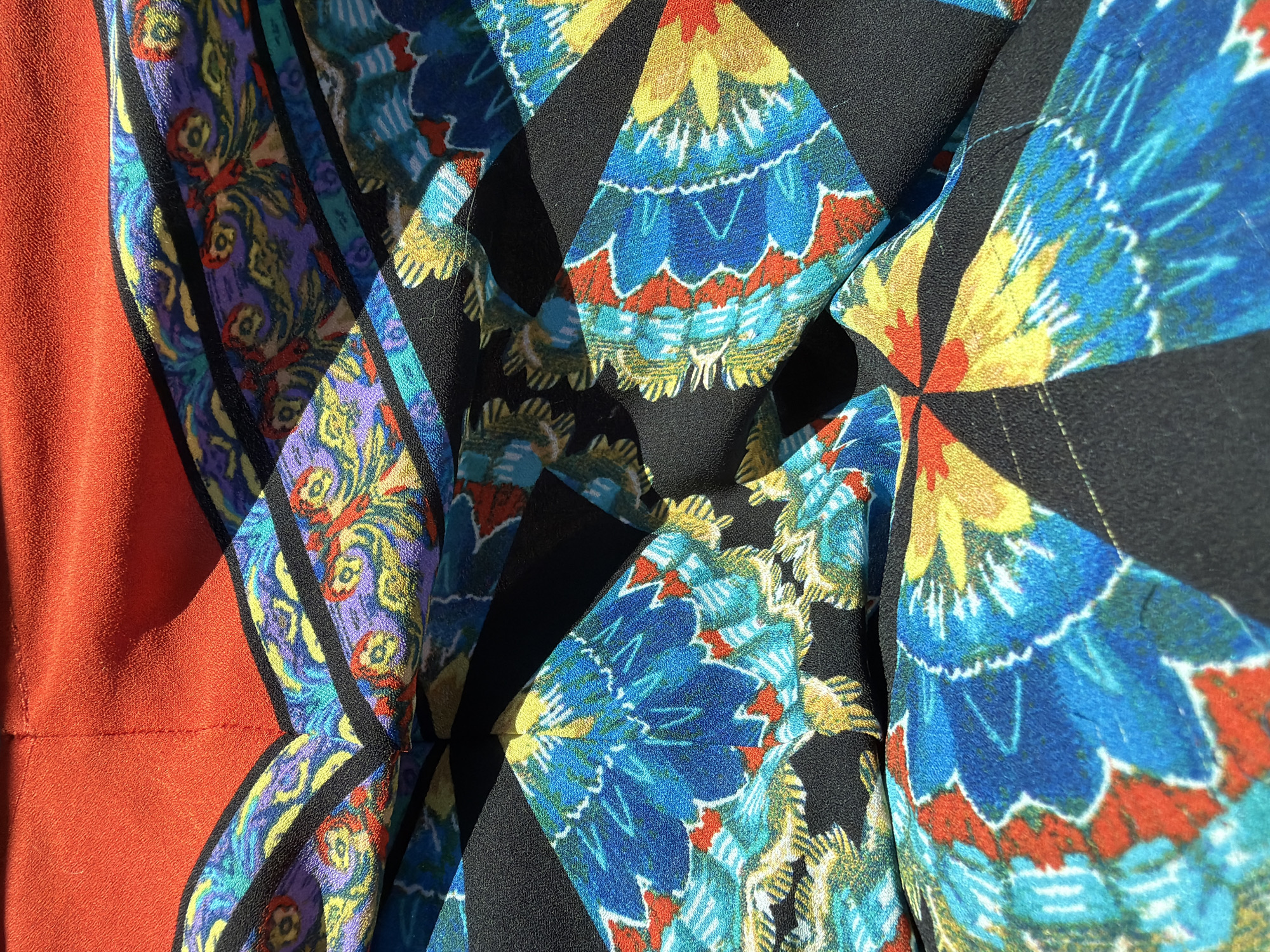 This is a close-up, overhead shot of brightly colored fabric. The fabric features a bold, abstract pattern with shapes resembling feathers or floral elements in shades of blue, yellow, orange, and purple. These colorful sections are interspersed with black fabric and bordered by black lines. The top portion of the image shows a solid section of bright red fabric. The fabric is creased and folded, creating shadows that add depth to the image. The location is Unknown, Unknown.