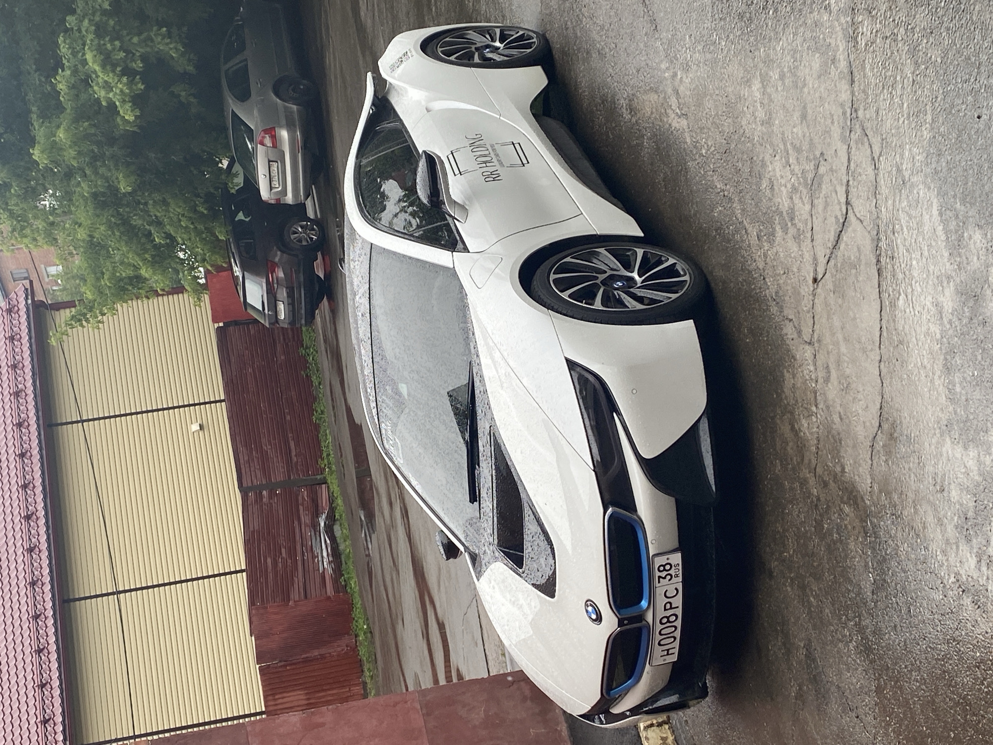 An outdoor medium shot captures a white BMW i8 parked on a wet asphalt surface. The car is angled, with its front facing towards the lower left of the frame. The vehicle exhibits a sleek, sporty design with distinctive blue accents on the grille. The license plate is clearly visible and reads "H008PC 38 RUS," indicating registration in the Irkutsk Oblast of Russia. The BMW's hood has a black inset, and the wheels are black with silver detailing.
To the left of the BMW, a portion of a dark red, corrugated metal fence is visible. Beyond the fence, a building with light yellow horizontal siding and a red tiled roof is present. In the background, two other vehicles are parked under a dense green tree. The rear of a dark-colored SUV is on the left, and a silver sedan is to its right. The license plate on the silver sedan appears to read "K376CV 735."
The ground is wet and shows cracks, with puddles reflecting the surrounding environment. The overall atmosphere suggests a cloudy or rainy day. There are no people visible in the image. The scene appears to be a street or a parking area in a residential or commercial district within the city of Irkutsk, Russia, based on the license plate information.